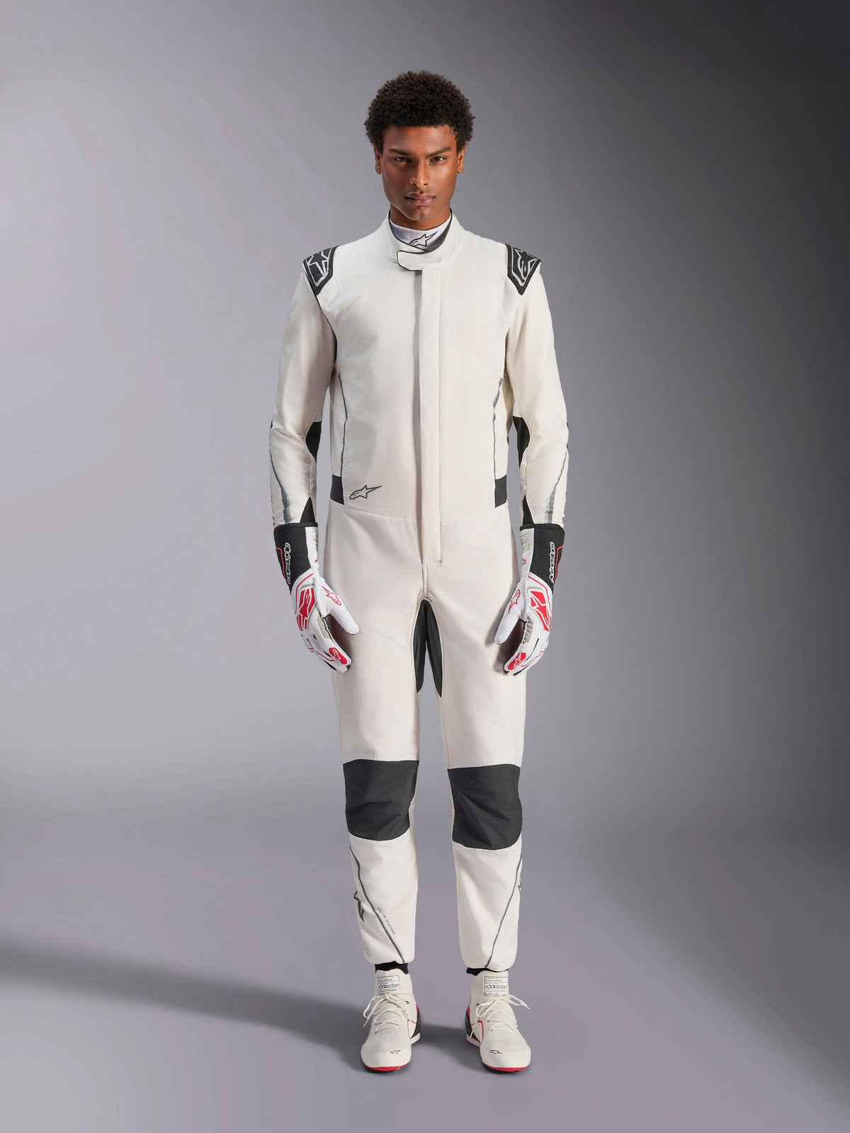 Alpinestars Hypertech V3 Suit, professioneller Autorennanzug, Weiß, Schwarz und Silber, getragen von einem Model mit rot-weißen Handschuhen und passenden weißen Rennschuhen, mit schwarzen Kontrasteinsätzen und Alpinestars-Logos, athletisches Slim-Fit-Design