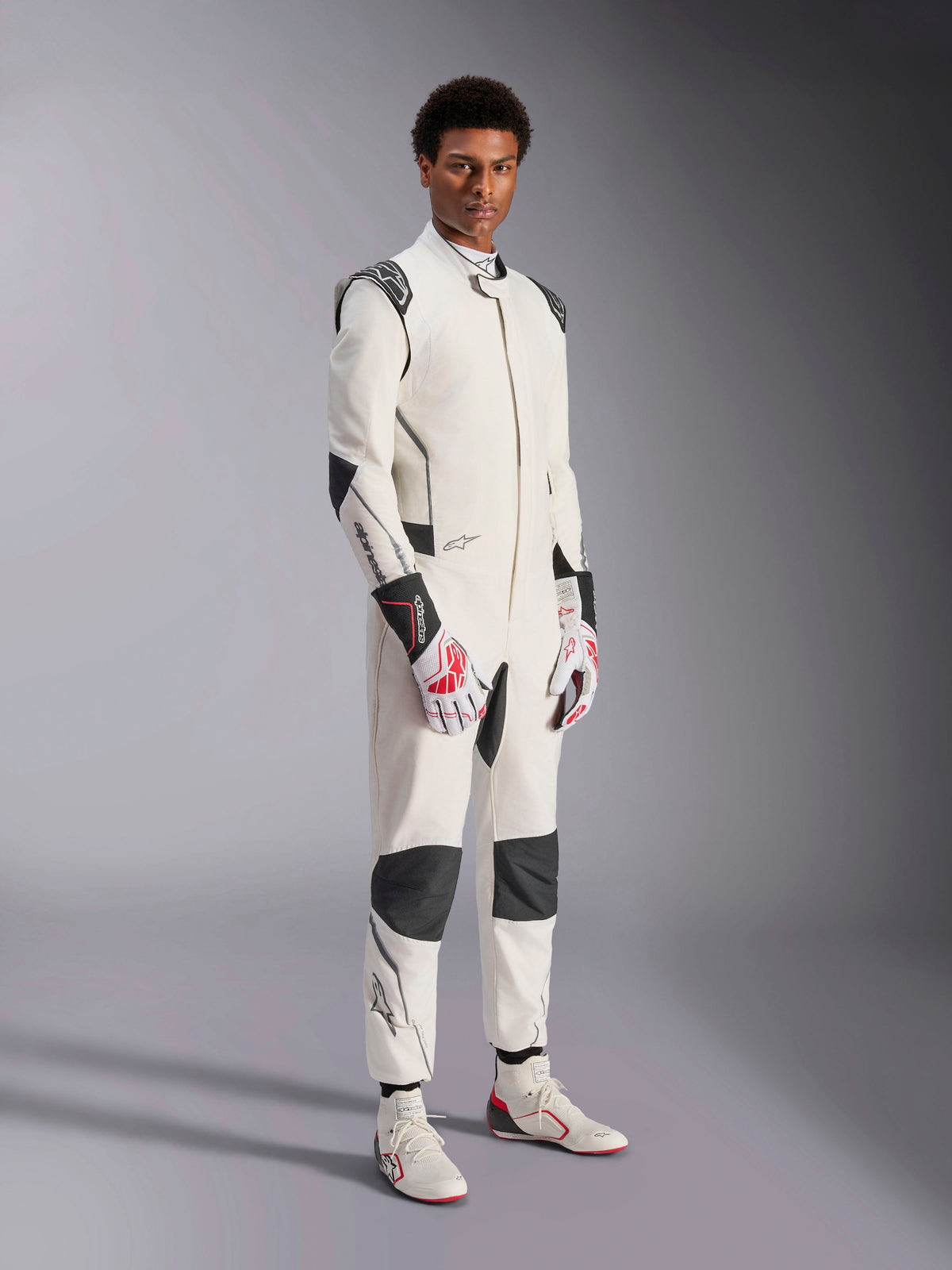 Männliches Model im Alpinestars Hypertech V3 Anzug, Weiß mit schwarzen und silbernen Akzenten, professionelle Automobilrennsport-Ausrüstung mit schwarzen Einsätzen an Schultern und Knien, kombiniert mit weiß-roten Rennhandschuhen und weißen Rennstiefeln