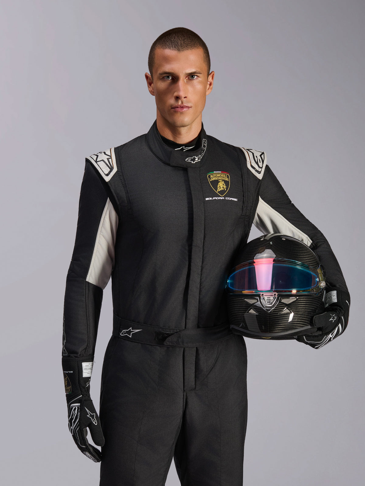 Männliches Model im Limited Edition Lamborghini GP Tech V4 Anzug, schwarzer Rennanzug mit silbernen Akzenten und Lamborghini Squadra Corse Logo, hält einen Carbonhelm, schwarze Rennhandschuhe mit weißen Logos, professionelle Autorennausrüstung