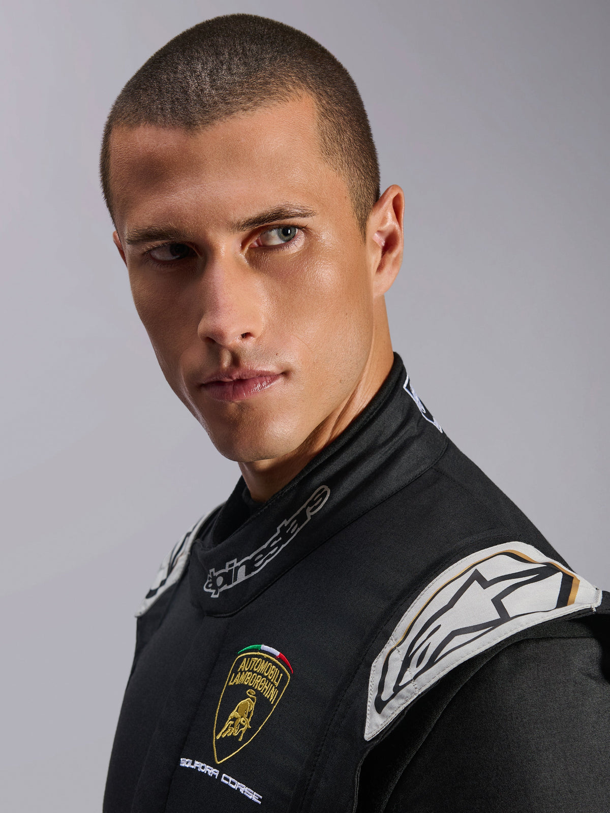 Nahaufnahme eines männlichen Models im Limited Edition Lamborghini GP Tech V4 Anzug in Schwarz, Silber und Gold, mit Logos von Automobili Lamborghini Squadra Corse und Alpinestars, professionelle FIA-zertifizierte Autorenn-Ausrüstung