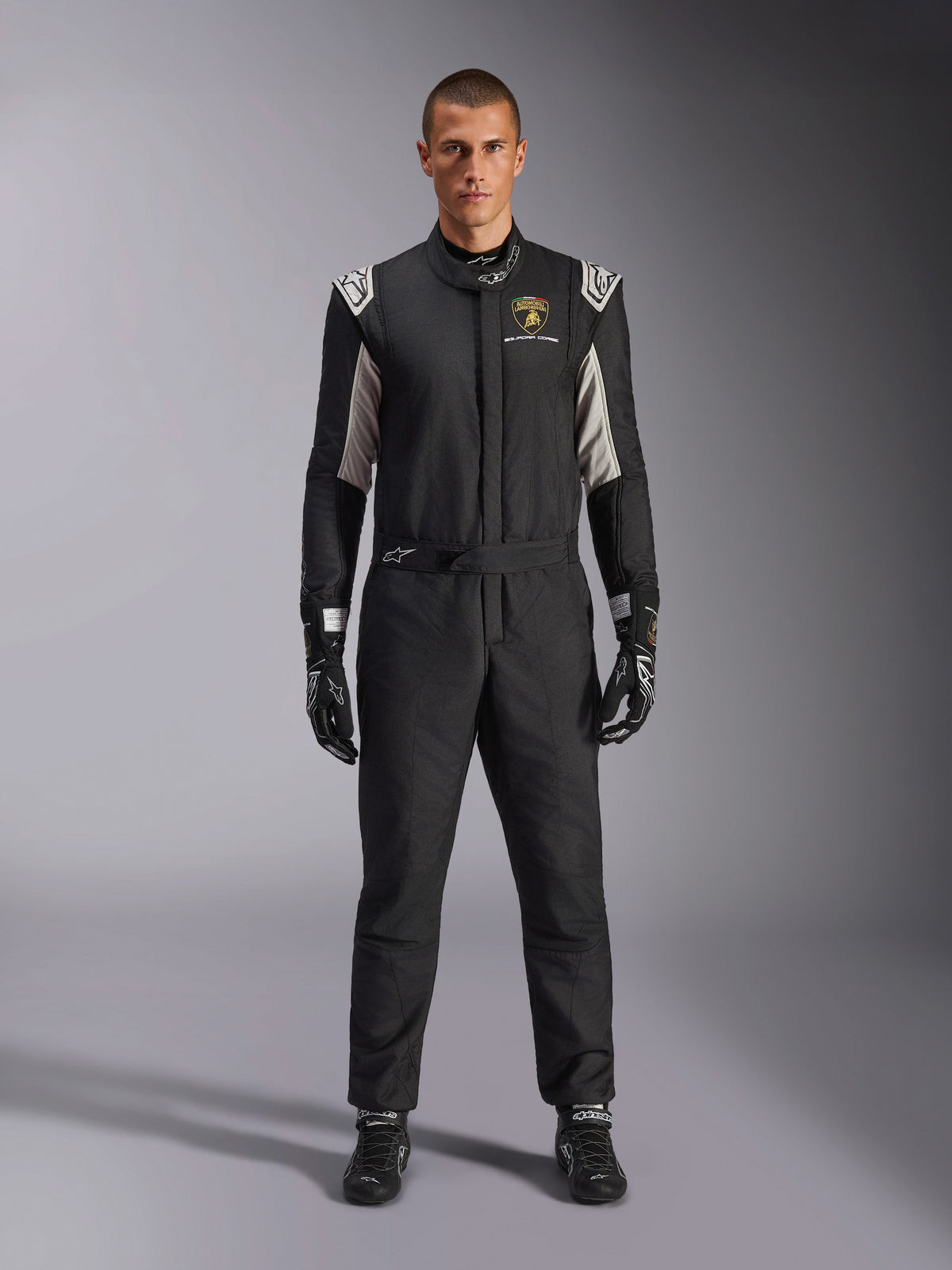 Alpinestars Limited Edition Lamborghini GP Tech V4 Anzug FIA, professioneller Autorennanzug, Schwarz mit silbernen und goldenen Akzenten, mit Automobili Lamborghini Squadra Corse Logo auf der Brust, getragen mit passenden Rennhandschuhen und Stiefeln, schlankes sportliches Design für Rundstreckenrennen