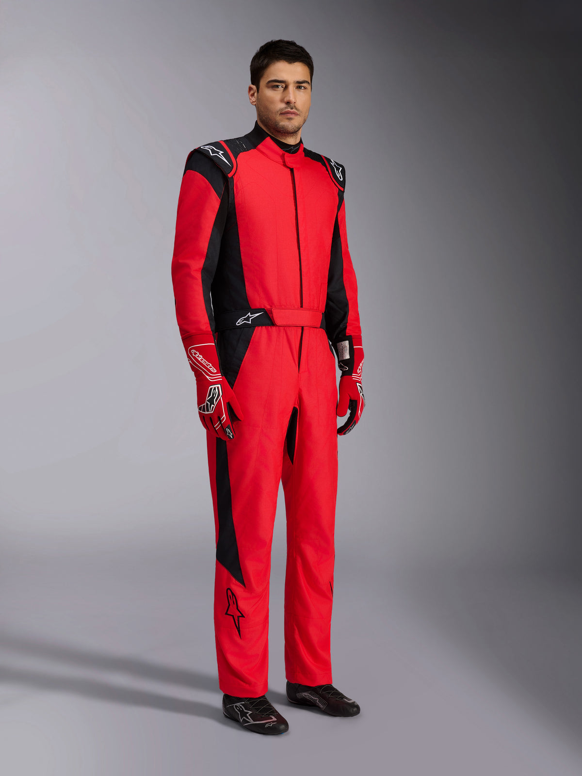 Alpinestars GP Pro Comp V2 FIA Racing Suit, Red and Black professional auto racing suit, worn with matching red and white gloves and black racing shoes, full-body studio portrait of motorsport protective gear.