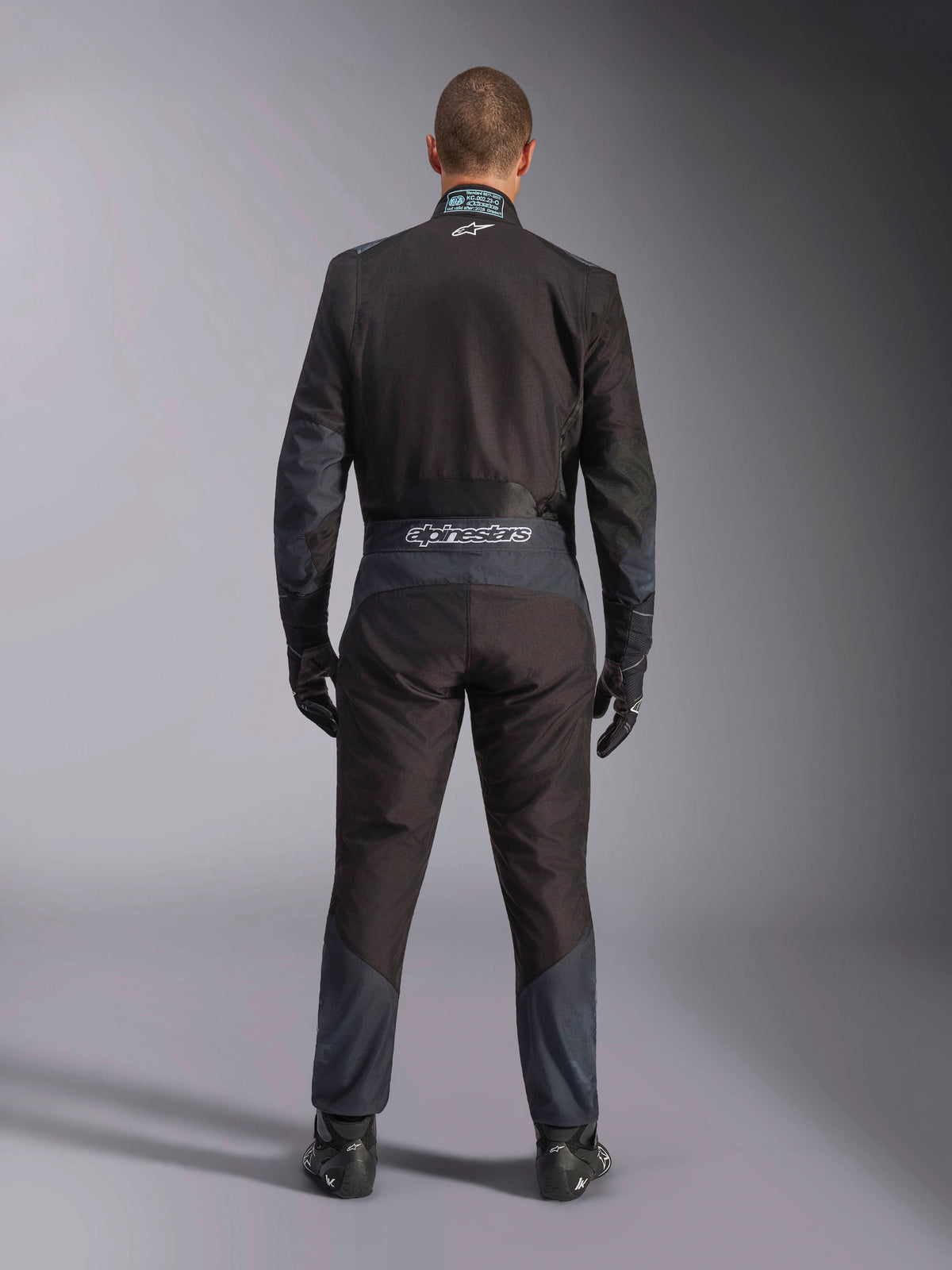 Alpinestars KMX-5 V3 Anzug, Kart-Anzug, Schwarz und Dark Shadow, Rückansicht eines Fahrers im kompletten Rennanzug mit passenden Handschuhen und Schuhen, mit Alpinestars-Branding am Hosenbund und ergonomischen Paneelen.