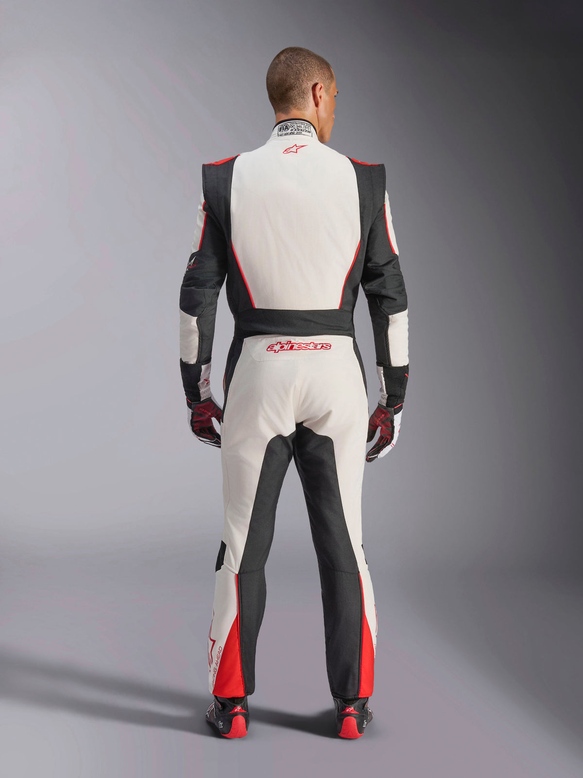Alpinestars GP Tech V4 Suit, Auto Racing Suit, White Black and Red, back view of driver wearing the one-piece suit, featuring black stretch panels on the sides and shoulders, red accents on the lower legs, Alpinestars branding on the lower back and collar, professional FIA-certified motorsport gear
