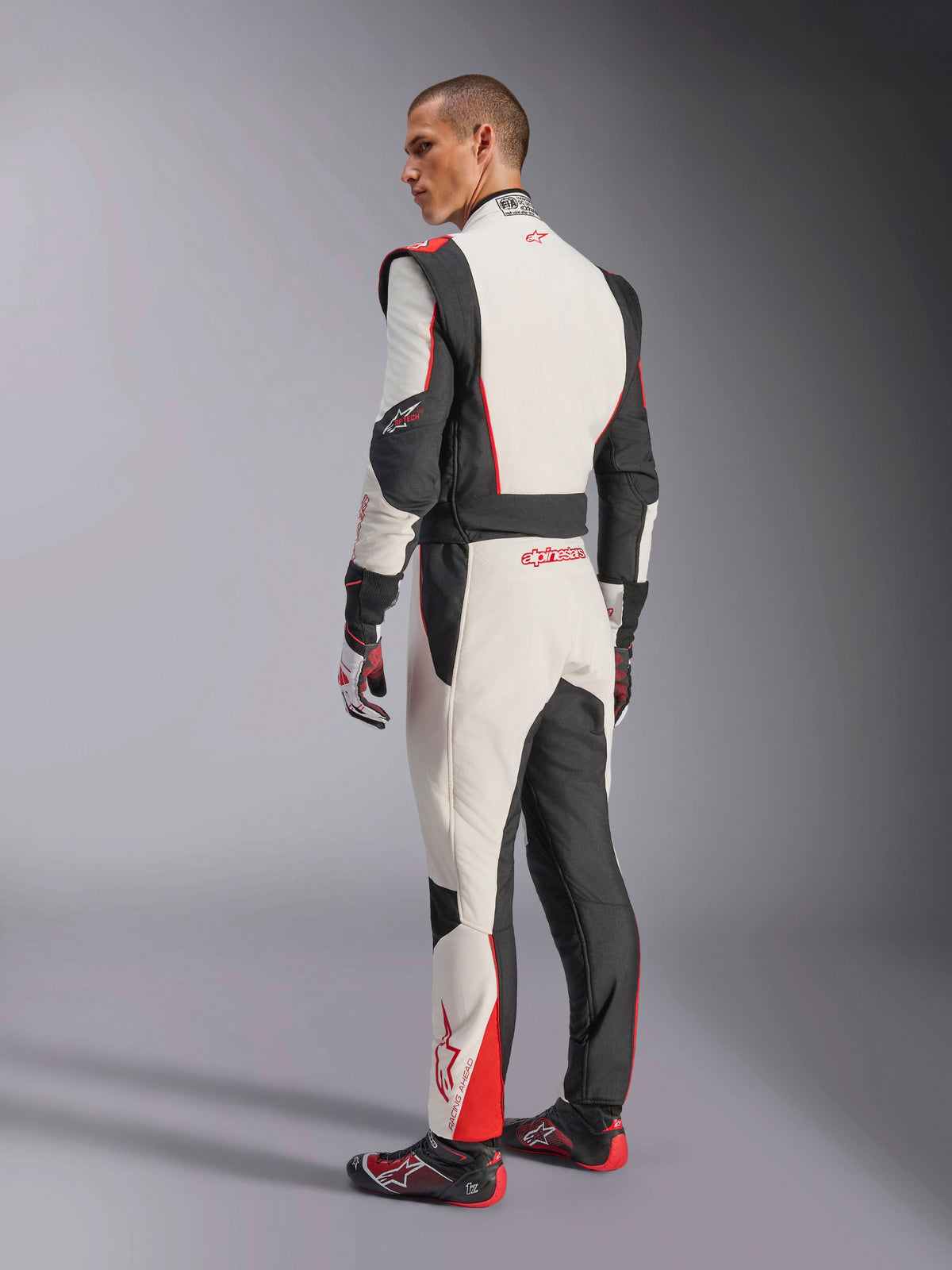 Alpinestars GP Tech V4 Suit, White Black Red racing suit, rear view showing ergonomic stretch panels on the lower back and legs, paired with matching racing gloves and shoes, professional motorsport apparel.