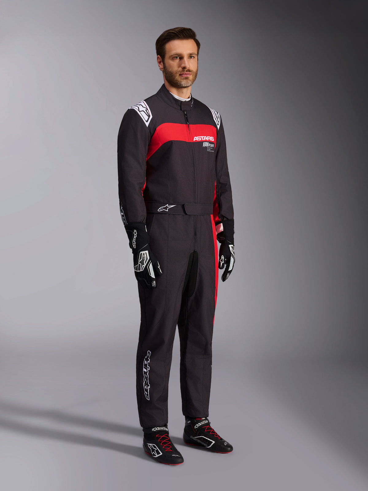 Männliches Modell im Alpinestars KMX-9 V3 Graphic 3 Anzug, Karting-Anzug, Schwarz und Rot, mit weißen Schulter-Epauletten und rotem Brust-Logo, kombiniert mit schwarzen Rennhandschuhen und Schuhen mit roten Schnürsenkeln, professionelle Motorsportbekleidung
