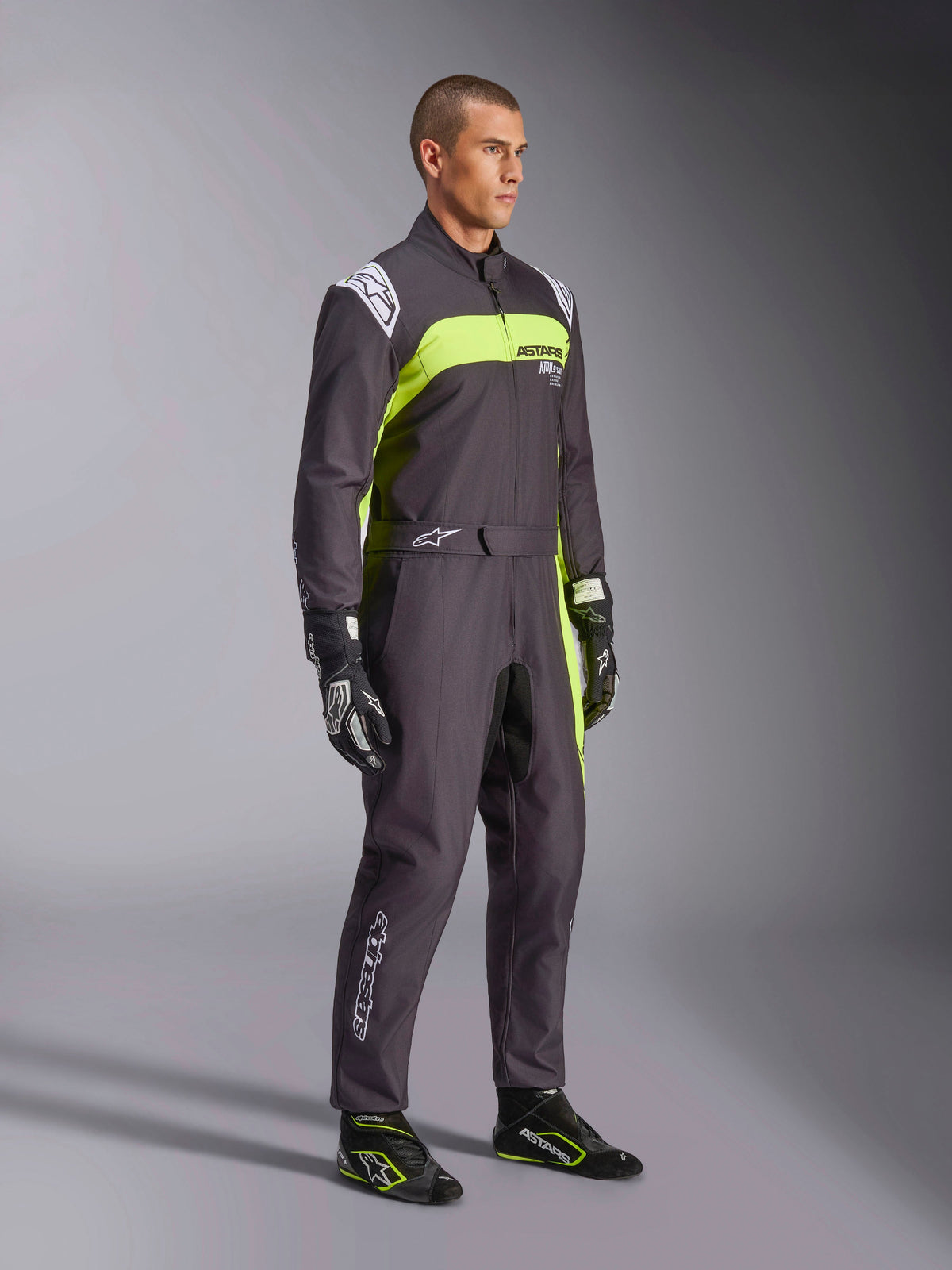 Männliches Model im Alpinestars KMX-9 V3 Graphic 3 Anzug in Schwarz und Gelb Fluo, professionelle Karting-Ausrüstung mit leuchtenden neongelben Einsätzen und weißen Logo-Akzenten, ergänzt durch farblich abgestimmte Rennhandschuhe und Schuhe, vor einem Studiohintergrund.