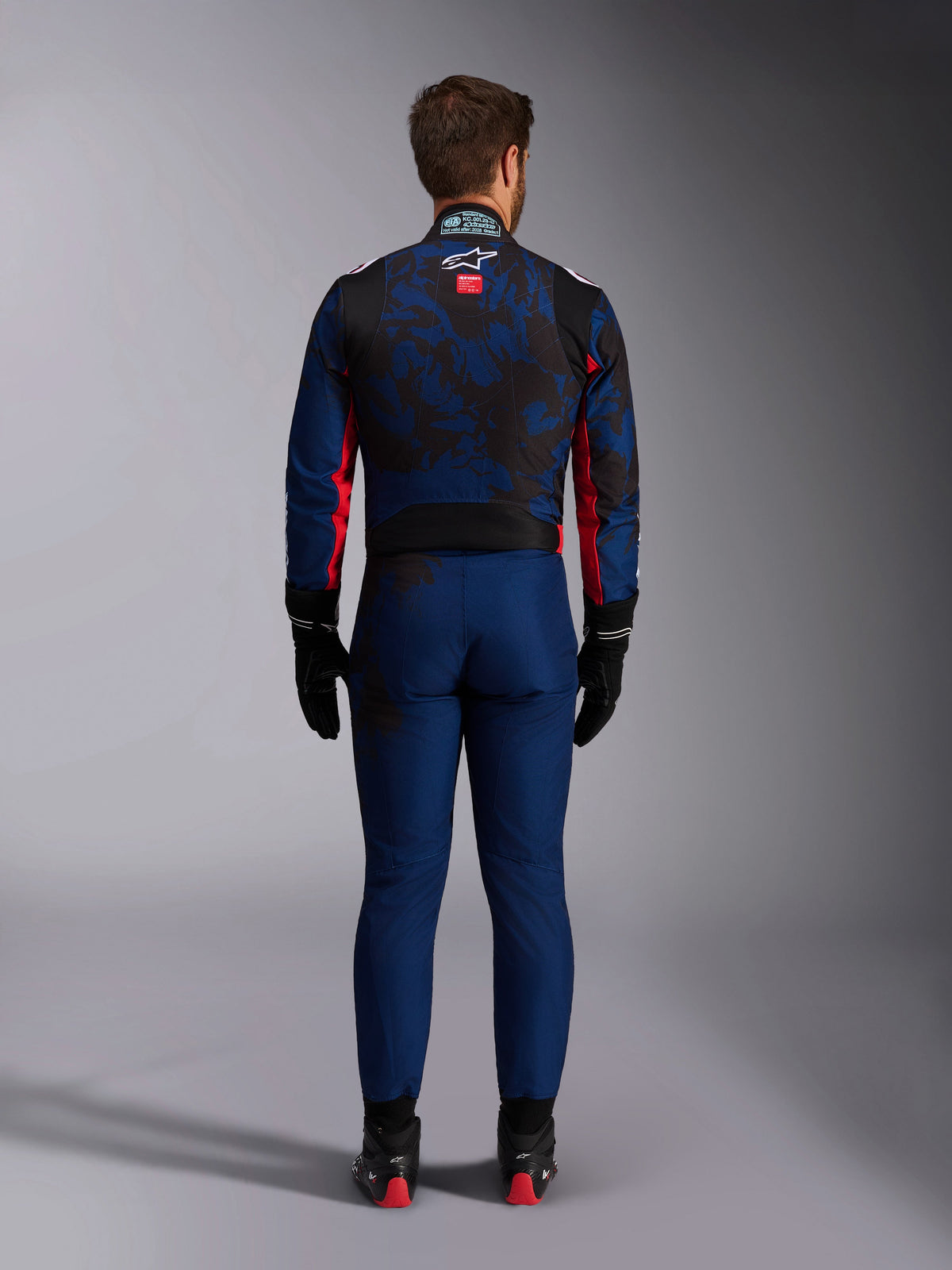 Kartfahrer in der Rückansicht im KMX-9 V3 Graphic 5 Anzug in Navy-Rot, mit einem navyblauen und schwarzen Grafikmuster, roten Seitenakzenten und dem Alpinestars-Logo auf dem oberen Rücken, kombiniert mit schwarzen Rennhandschuhen und Schuhen.