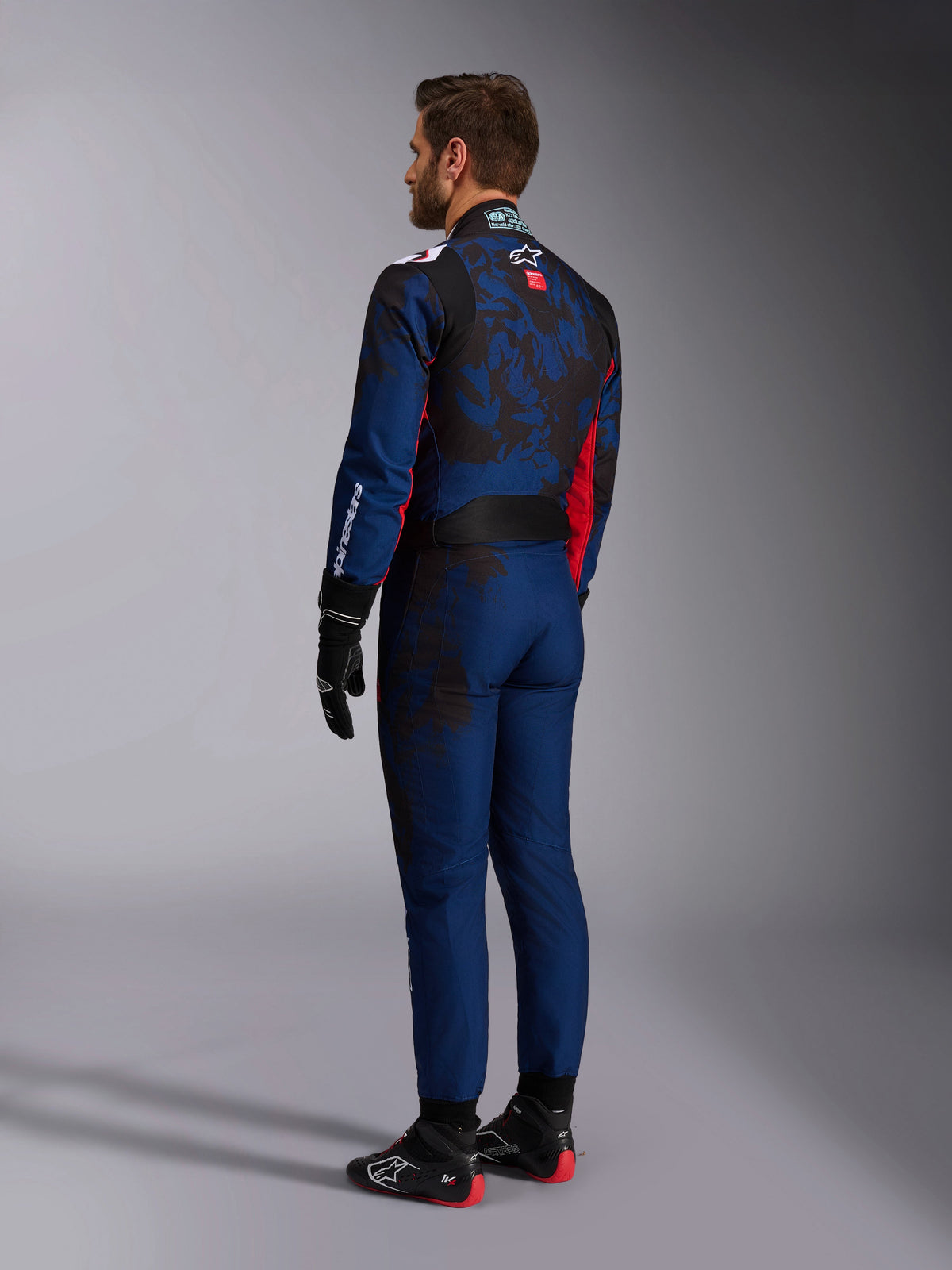 Alpinestars KMX-9 V3 Graphic 5 Anzug, Kart-Anzug, Navy-Rot, Rückansicht mit einem navyblauen Hauptteil, einer dunklen Camouflage-Grafik auf dem Rücken und roten Akzenten, getragen mit schwarzen Rennhandschuhen und schwarz-roten Kartschuhen.