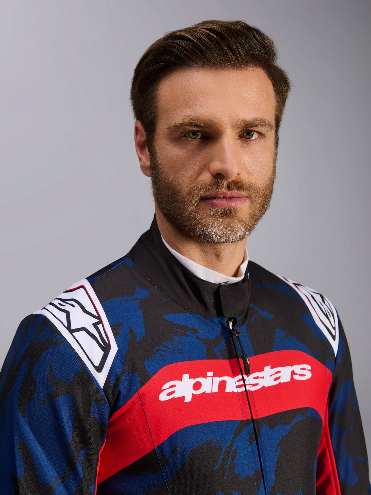 Alpinestars KMX-9 V3 Graphic 5 Anzug, Kart-Anzug in Navy-Rot, mit blau-schwarzem Camouflage-Grafikmuster, auffälligem rotem Querstreifen mit weißem Alpinestars-Schriftzug auf der Brust und weißen Logos auf den Schultern, getragen von einem Model in einer Studioumgebung.