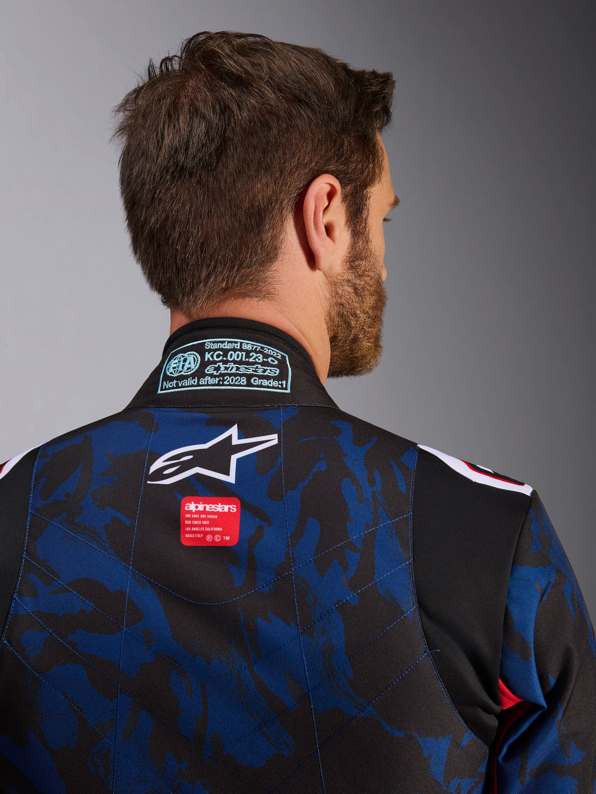 Nahaufnahme des Alpinestars KMX-9 V3 Graphic 5 Anzugs, Kart-Anzug in Navy-Rot, mit blau-schwarzem Camouflage-Muster, weißem Stern-Logo und rotem Markenlabel auf dem oberen Rücken, mit FIA 8877-2022 Zertifizierungsaufnäher am Kragen