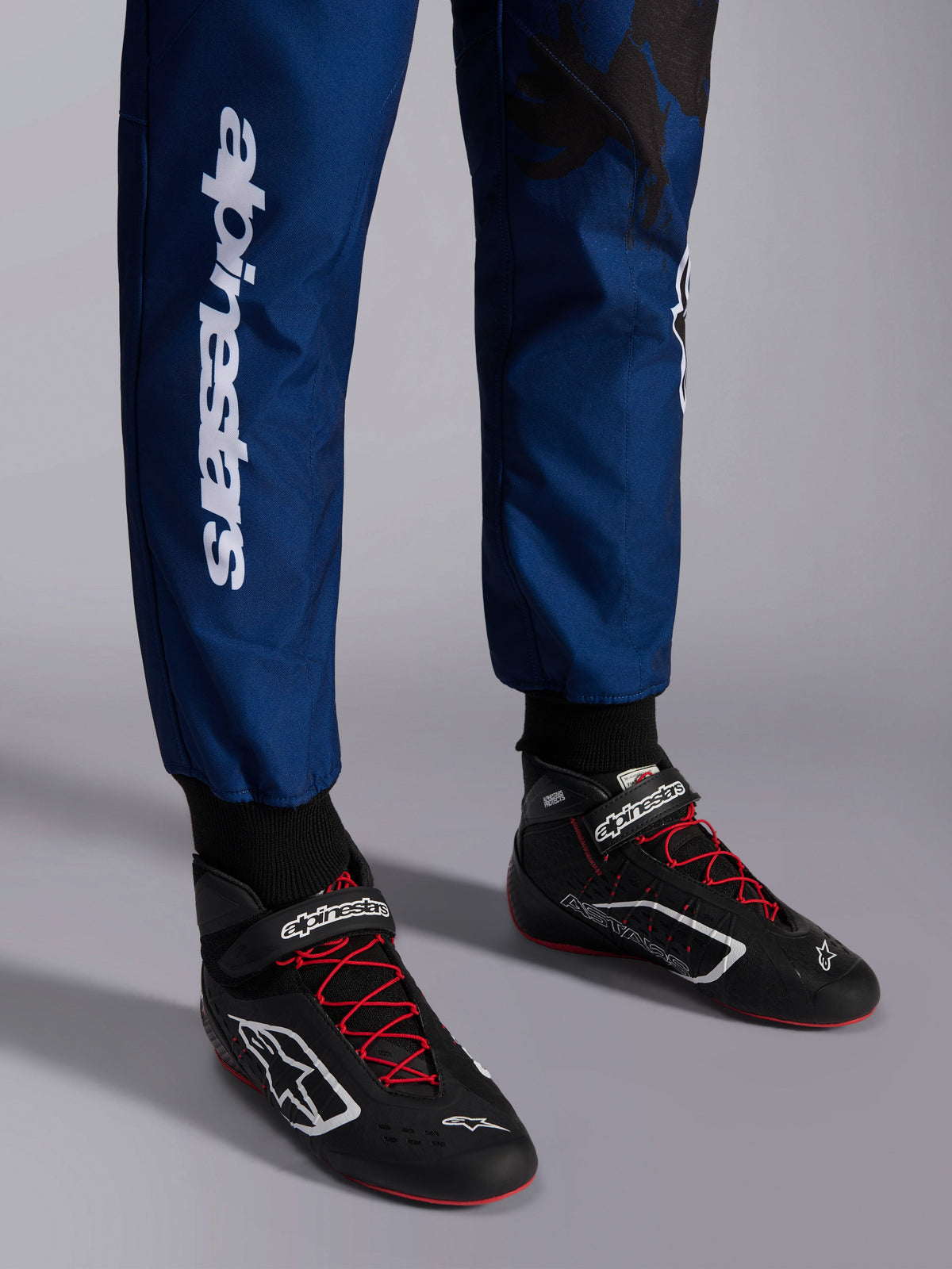 Alpinestars KMX-9 V3 Graphic 5 Anzug, Navy-Rot, Nahaufnahme der Unterschenkel und Kart-Schuhe, dunkelblaues Material mit weißem vertikalem Alpinestars-Schriftzug, schwarze Schuhe mit roten Schnürsenkeln und weißen Logos, professionelle Kart-Ausrüstung