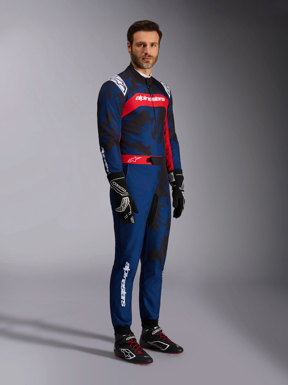 Model im Alpinestars KMX-9 V3 Graphic 5 Anzug in Navy Rot, Ganzkörperaufnahme mit schwarzen Kart-Handschuhen und schwarzen Rennschuhen, marineblauer Anzug mit dunklen Camo-Grafiken und auffälligem roten Brustpanel mit weißem Branding, professionelle Kart-Schutzausrüstung