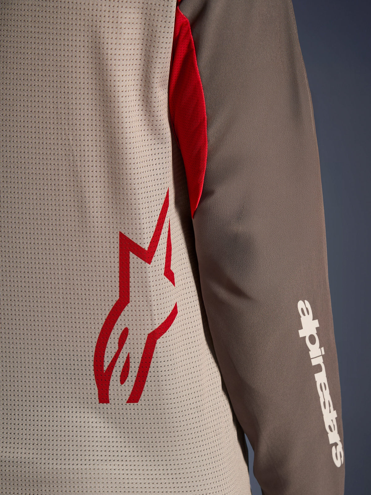 Alpinestars Maxdura Dual Jersey, Aluminum Walnut, Nahaufnahme des belüfteten Mesh-Torsos mit einem roten Stern-Logo und einem braunen, abriebfesten Ärmel mit weißem Alpinestars-Schriftzug, technische Adventure- und Dual-Sport-Motorradbekleidung