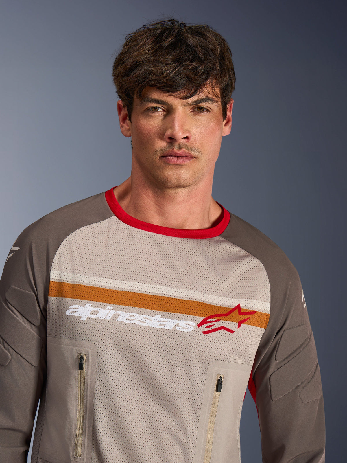 Männliches Model trägt Alpinestars Maxdura Dual Jersey in Aluminum Walnut, Adventure-Motorradtrikot mit Rumpfbereich aus vollständigem Mesh, abriebfesten Ärmeln, rotem Kragen und Brusttaschen mit Reißverschluss, mit orange-weißen Bruststreifen und Alpinestars-Logo.