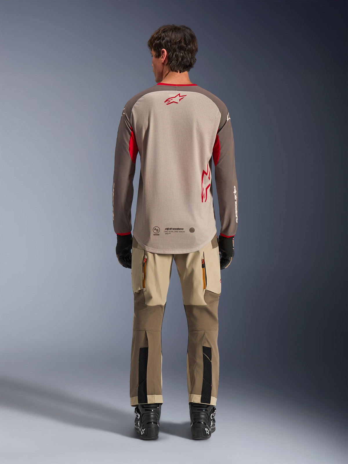 Alpinestars Maxdura Dual Jersey in Aluminum Walnut, Rückansicht, beigefarbener Mesh-Torso mit braunen Ärmeln und roten Logos, verlängerter Rückensaum, kombiniert mit Adventure-Fahrhose und schwarzen Stiefeln.
