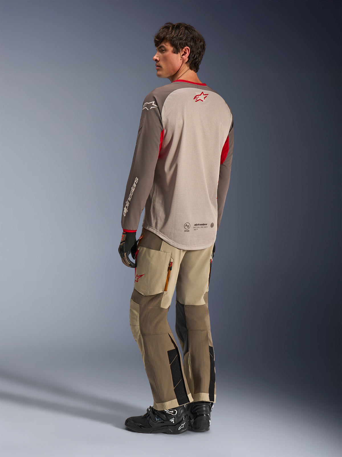 Alpinestars Maxdura Dual Jersey in Aluminum Walnut, Rückansicht eines Adventure-Motorradtrikots mit atmungsaktivem Mesh-Torso und roten Akzenten, kombiniert mit passender technischer Adventure-Hose und schwarzen Stiefeln