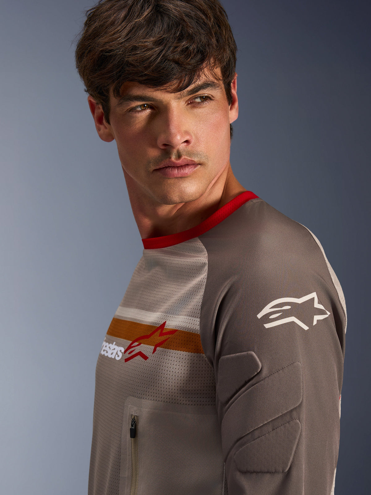 Männliches Model im Alpinestars Maxdura Dual Jersey in Aluminum Walnut, mit belüftetem Mesh-Torso, rotem Kragen und orangem Bruststreifen. Nahaufnahme des abriebfesten Ärmels mit integrierter Polsterung und einer Reißverschluss-Brusttasche für Adventure-Fahrten.