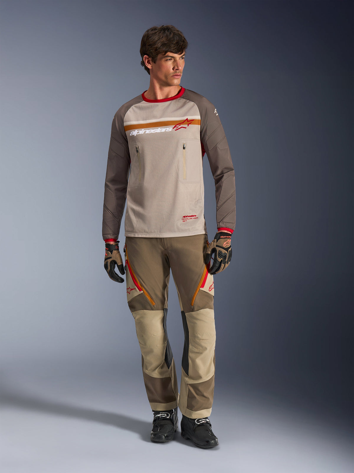 Alpinestars Maxdura Dual Trikot in Aluminum Walnut, Adventure-Motorradtrikot mit beigem Mesh-Torso und braunen Ärmeln, mit rotem Kragen und horizontalem Bruststreifen mit Alpinestars-Logo, getragen mit passender Adventure-Hose und Handschuhen.