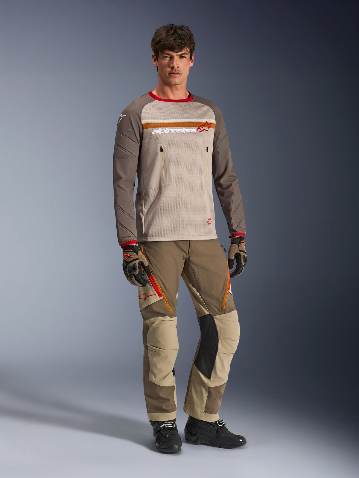 Model trägt das Alpinestars Maxdura Dual Jersey in Aluminum Walnut, mit einem grauen Mesh-Torso, braunen Ärmeln mit roten Bündchen und Brusttaschen mit Reißverschluss, kombiniert mit passender Offroad-Ausrüstung für Adventure-Fahrten.