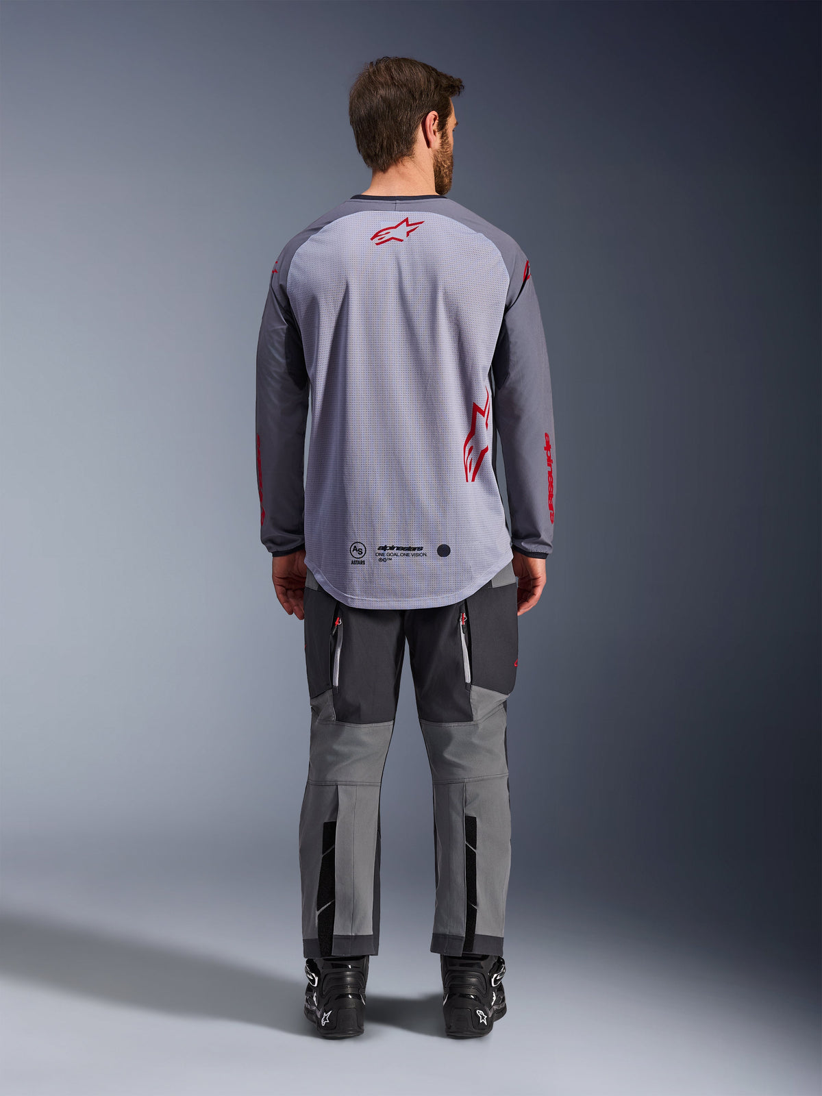 Männliches Model in der Rückansicht trägt das Alpinestars Maxdura Dual Jersey, Adventure-Trikot in Mittelgrau und Dunkelgrau mit roten Logo-Akzenten, Mesh-Rückenkonstruktion, kombiniert mit passender grauer Offroad-Hose und schwarzen Stiefeln
