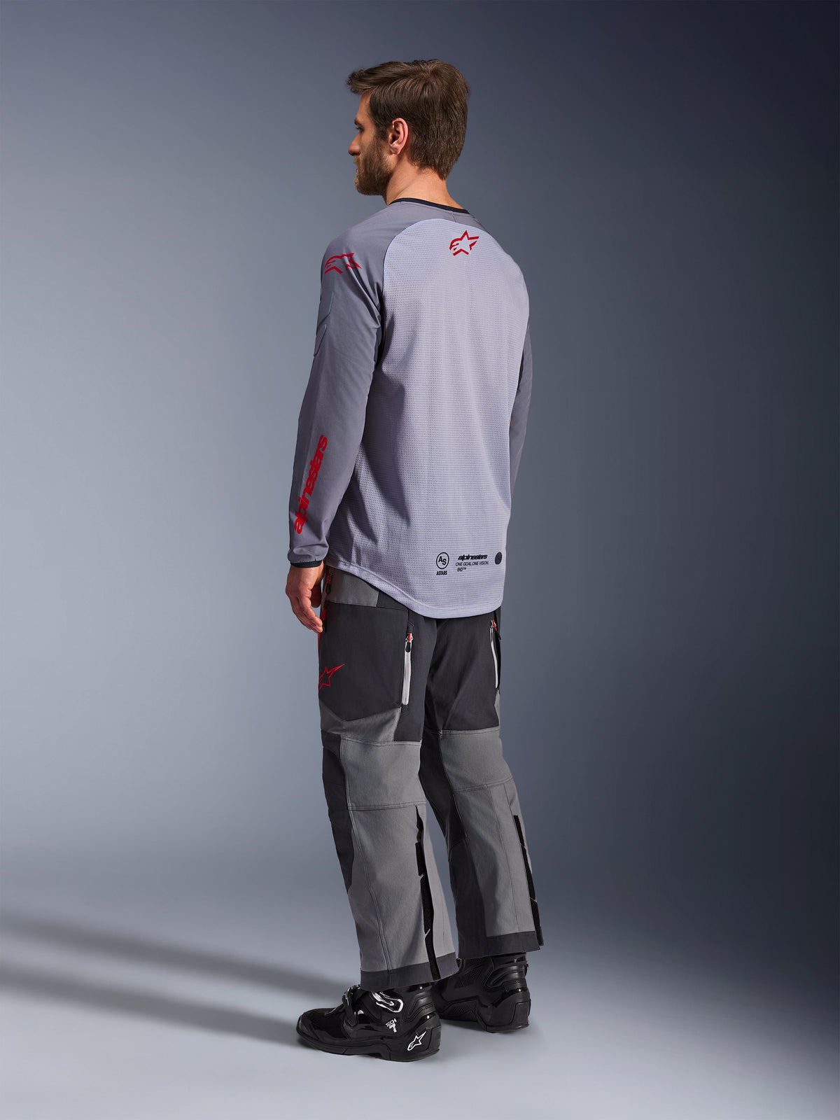 Mann im Alpinestars Maxdura Dual Jersey, hellgrau mit roten Alpinestars-Logos, Rückansicht mit Mesh-Torso und langen Ärmeln, kombiniert mit anthrazitfarbener technischer Hose und schwarzen Motorradstiefeln