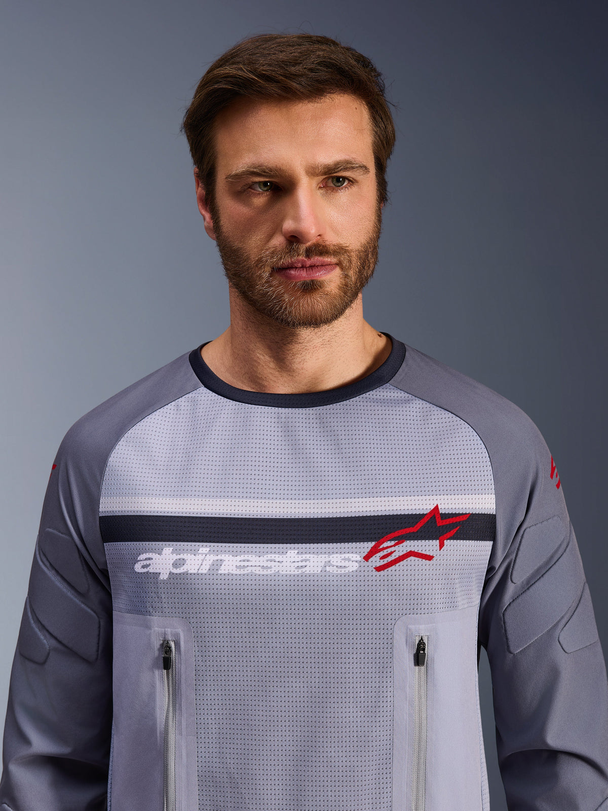 Alpinestars Maxdura Dual Jersey, Mittelgrau und Dunkelgrau, getragen von einem männlichen Model, Mesh-Torso für Belüftung, zwei Brusttaschen mit Reißverschluss, weißes „alpinestars“-Branding und rotes Stern-Logo, Performance-Offroad-Adventure-Ausrüstung