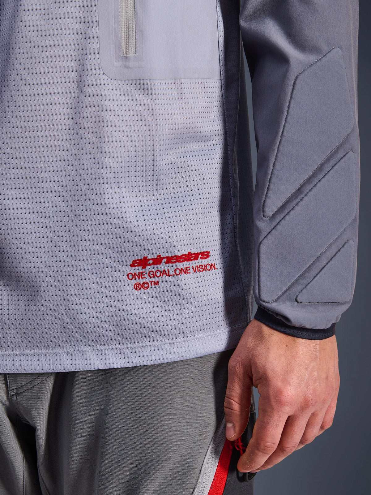 Alpinestars Maxdura Dual Jersey, Mid Gray und Dark Gray, Nahaufnahme des atmungsaktiven Mesh-Torsos mit rotem Branding, Reißverschlusstaschen-Detail und integrierter Polsterung am Ärmel, technische Adventure-Motorradausrüstung