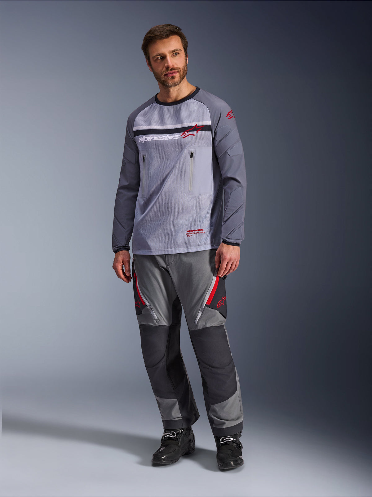 Ein Mann trägt das Alpinestars Maxdura Dual Jersey in Mittelgrau und Dunkelgrau, kombiniert mit einer farblich abgestimmten grau-schwarzen Adventure-Hose und schwarzen Stiefeln, mit atmungsaktivem Mesh-Torso, Brusttaschen mit Reißverschluss und roten Logo-Akzenten.