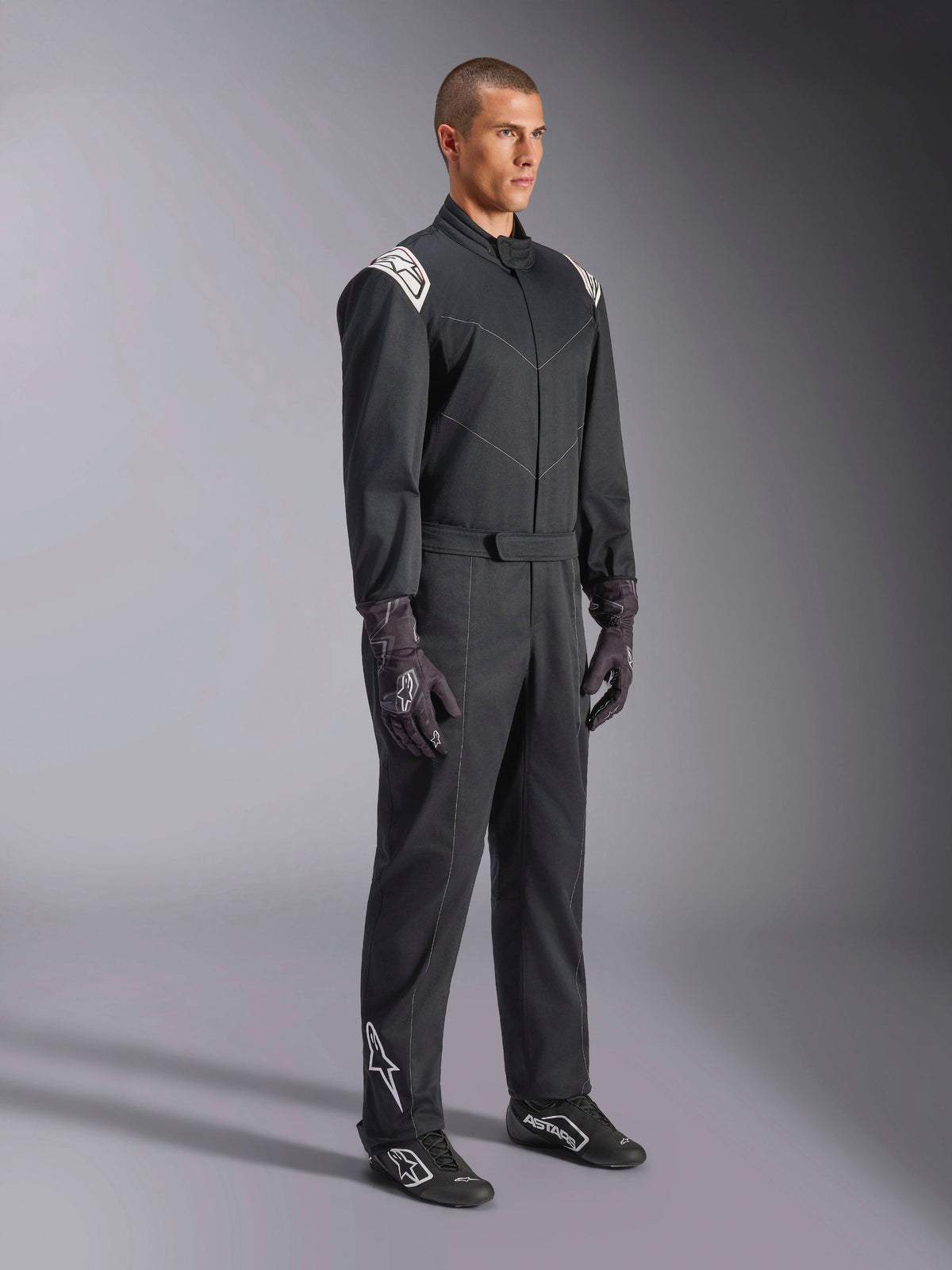 Alpinestars Kart Indoor V2 Suit, schwarzer einteiliger Kart-Anzug, von einem Model mit Rennhandschuhen und Schuhen getragen, weiße Schulterakzente und weißes Astars-Logo am Unterschenkel, professionelle Motorsportbekleidung für den Indoor-Einsatz