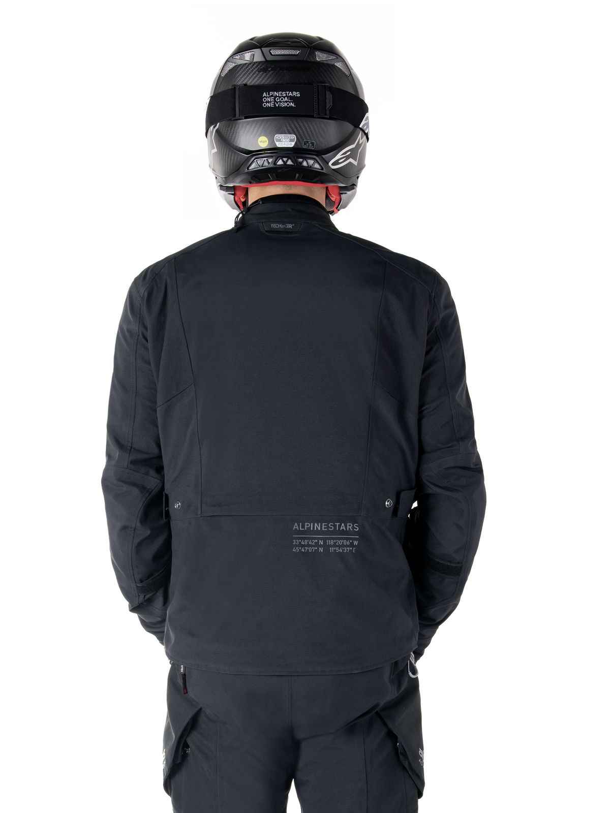 Alpinestars St-7 Prime GORE-TEX Jacke, Schwarz, Rückansicht eines Fahrers, mit dezentem Logo und geografischen Koordinaten auf dem unteren Rücken, getragen mit passendem schwarzem Helm und Hose, elegante und technische Motorradbekleidung
