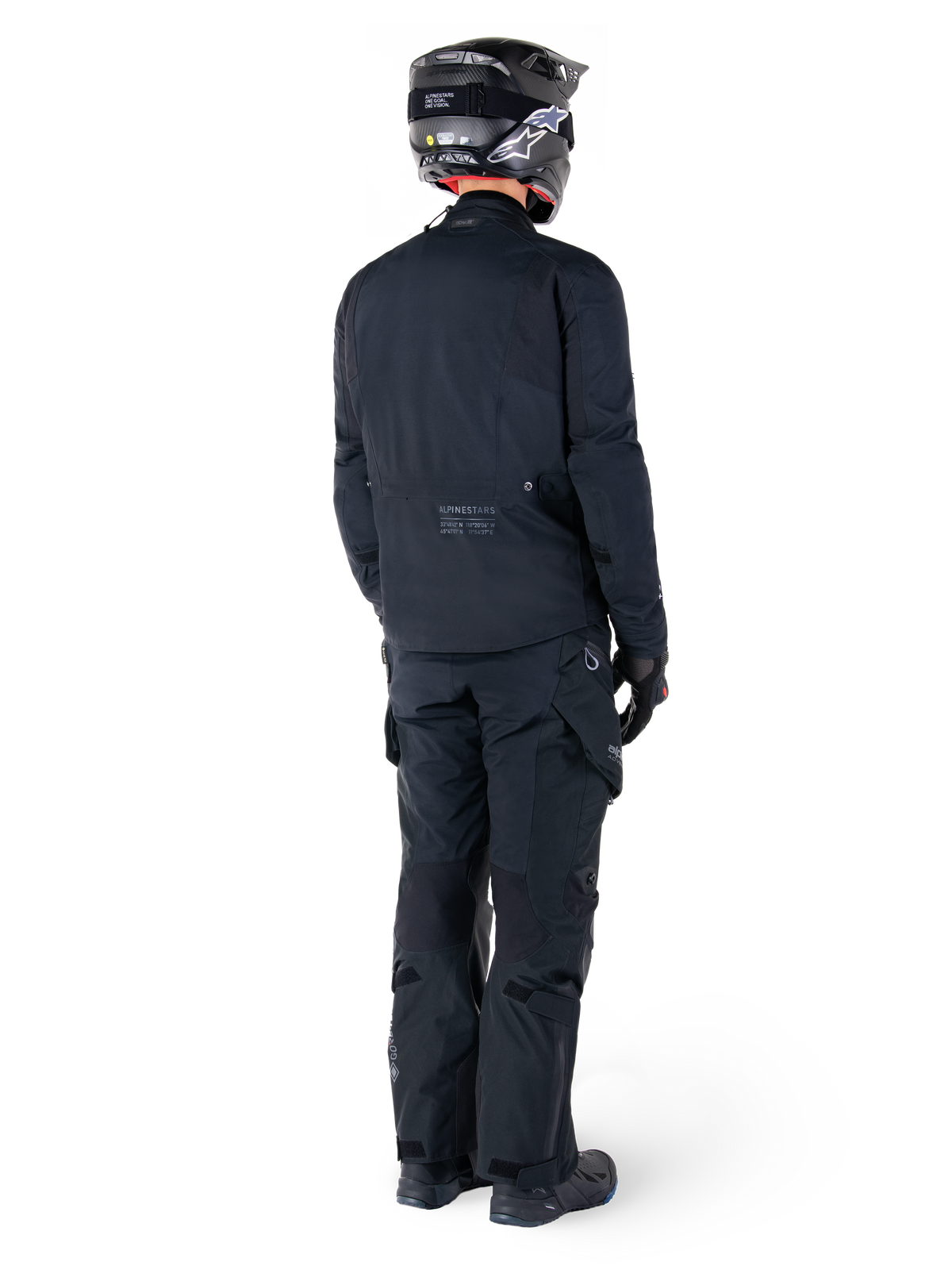 Rückansicht eines Fahrers mit der Alpinestars St-7 Prime GORE-TEX Jacke in Schwarz, kombiniert mit einer passenden schwarzen Tourenhose, einem schwarzen Motorradhelm mit weißem Logo und schwarzen Motorradstiefeln, komplette Adventure-Schutzausrüstung für Touren und Pendelfahrten.