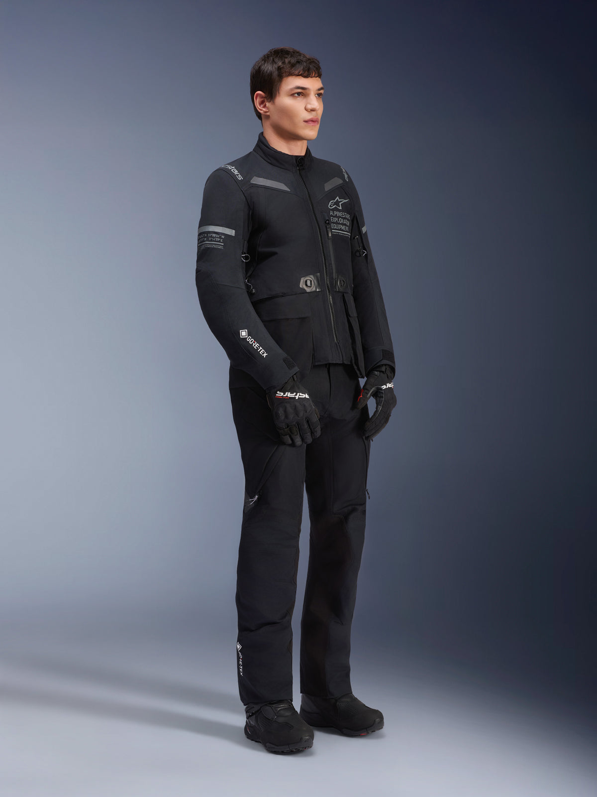 Alpinestars ST-7 2L GORE-TEX Hose, getragen von einem Modell mit der passenden ST-7 Jacke, komplett schwarzes Adventure-Touring-Motorrad-Outfit, mit GORE-TEX-Branding, Cargo-Taschen sowie schwarzen Handschuhen und Stiefeln, Studioaufnahme vor grauem Hintergrund