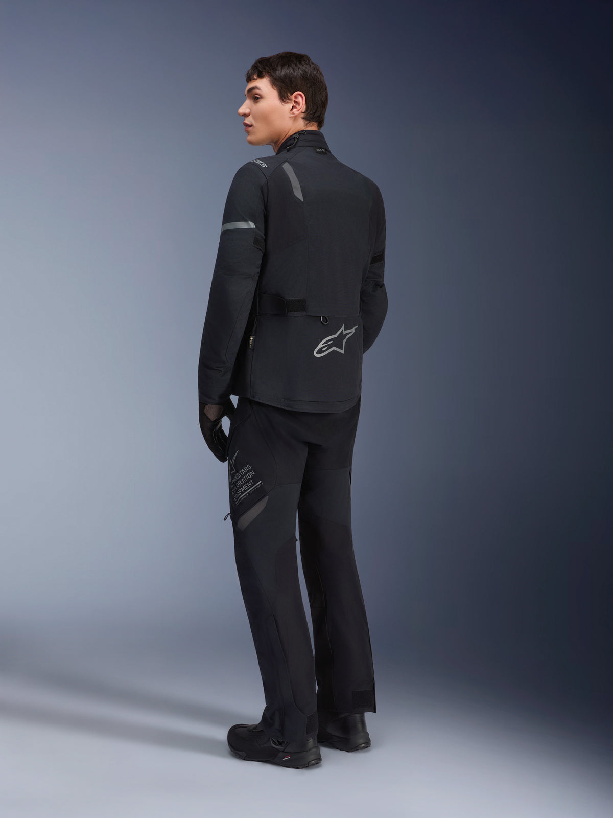 Alpinestars ST-7 2L GORE-TEX Hose, Adventure-Tourenhose, Schwarz und Dunkelgrau, getragen mit passender ST-7 Jacke und schwarzen Stiefeln, mit „Alpinestars Exploration Equipment“-Branding auf dem Oberschenkel, konzipiert für wasserdichtes und atmungsaktives Fahren in allen vier Jahreszeiten
