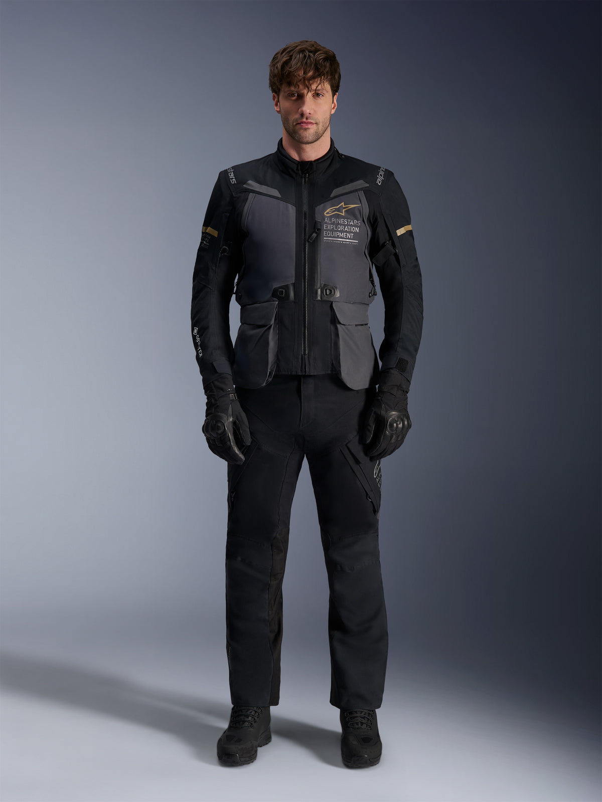 Männliches Model trägt die Alpinestars ST-7 2L GORE-TEX Jacke in Deep Gray Black Dark Khaki, mit dem „ALPINESTARS EXPLORATION EQUIPMENT“-Branding auf der Brust, kombiniert mit passender schwarzer Motorradhose, Handschuhen und Stiefeln, Studio-Frontansicht.
