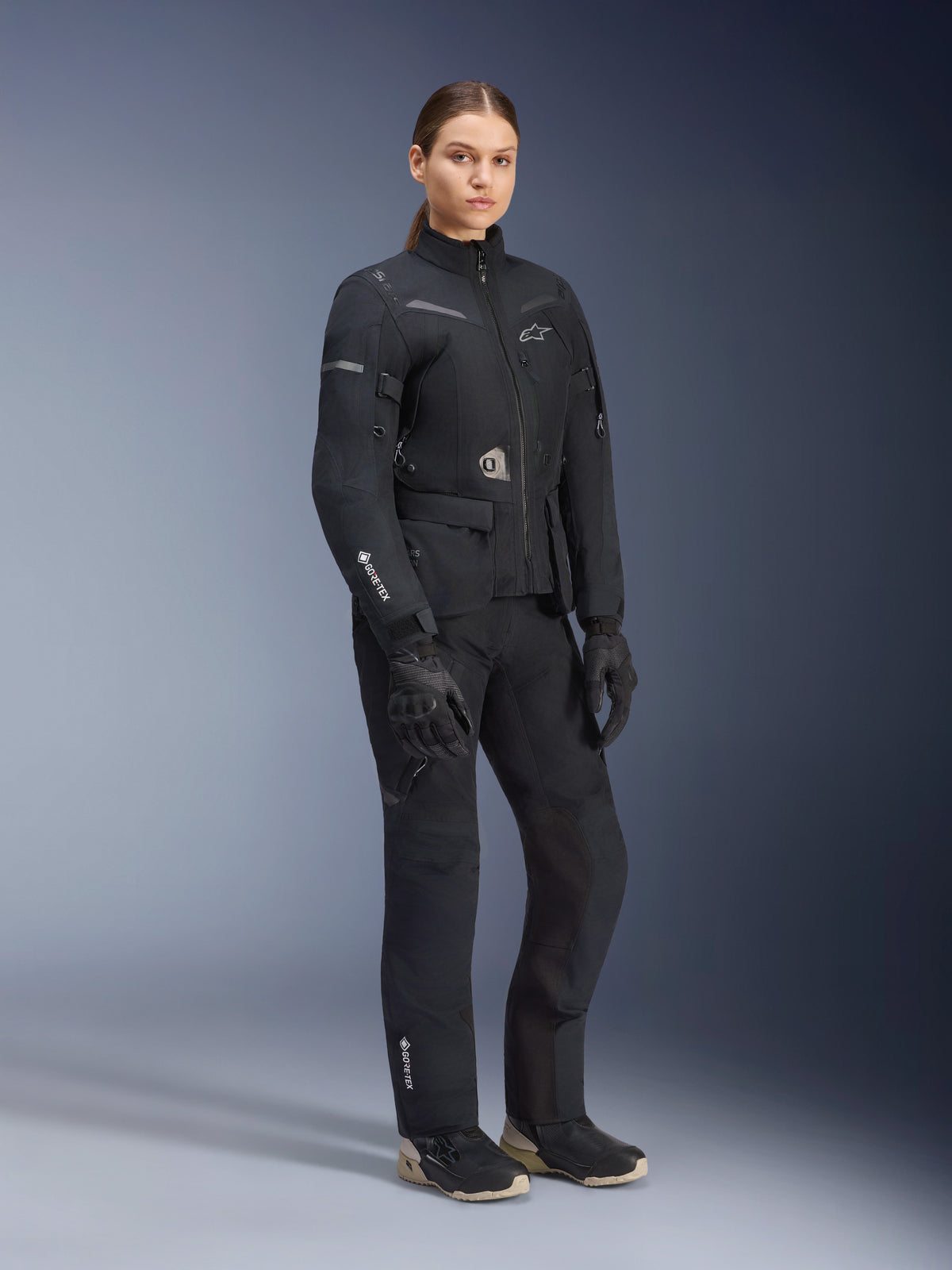 Woman model wearing Alpinestars Stella ST-7 2L GORE-TEX adventure touring suit, Black and Dark Gray jacket and pants, featuring prominent GORE-TEX branding on the sleeve and leg, paired with black gloves and riding boots, professional motorcycle gear designed for all-weather touring.