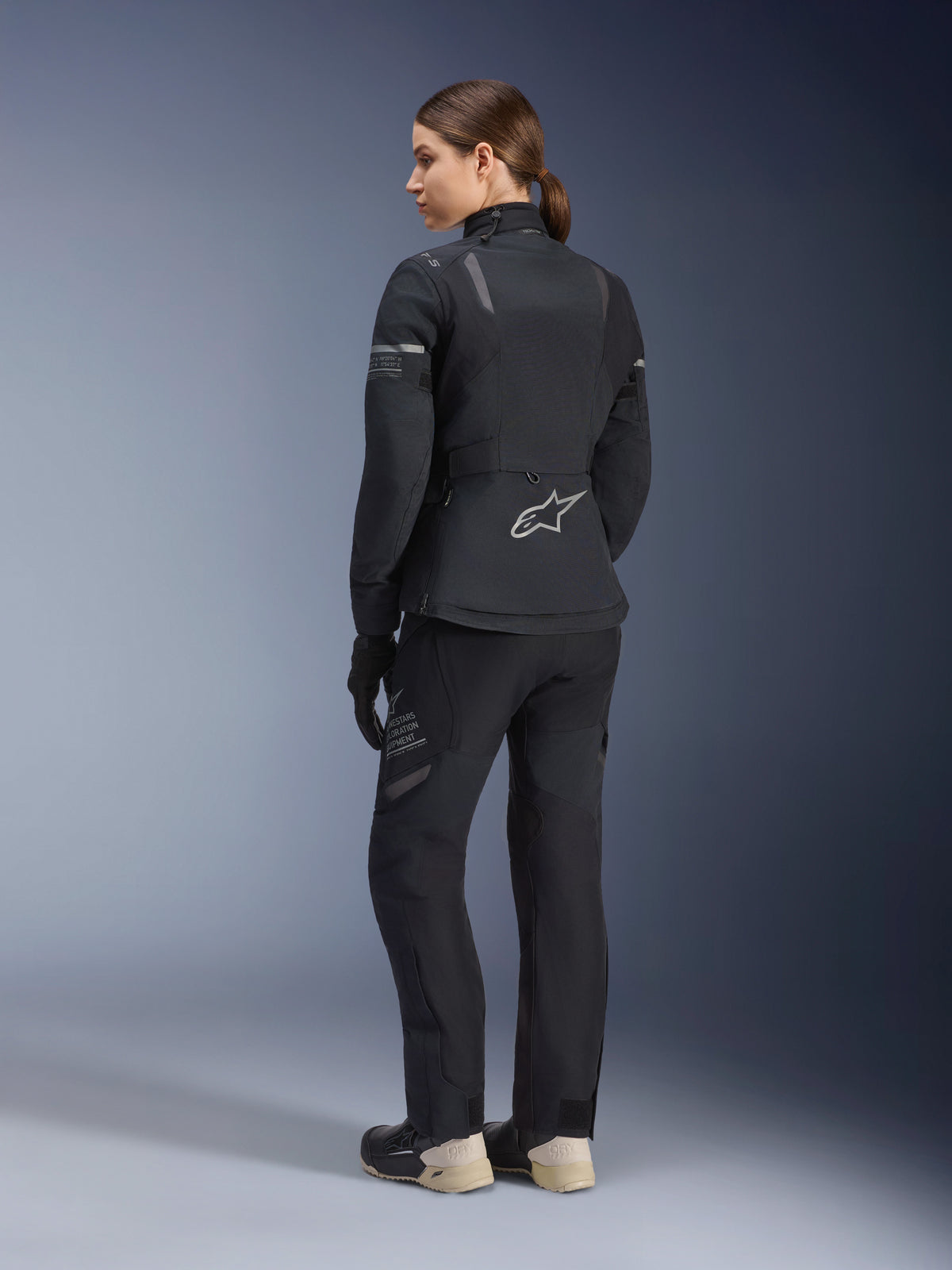 Alpinestars Women's Stella ST-7 2L GORE-TEX Jacket and Pants, full black adventure touring suit, back view showing Alpinestars star logo and Exploration Equipment branding, paired with beige boots and black gloves.