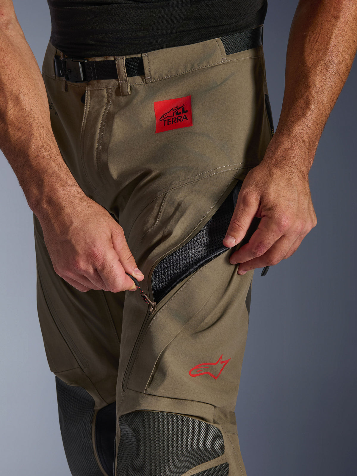 Alpinestars Nazca 3L GORE-TEX PRO Pants in Walnut Black, close-up detail of the thigh area showing a hand opening the large zippered ventilation intake, featuring a red ALL TERRA patch and red Alpinestars logo, technical adventure motorcycle gear.