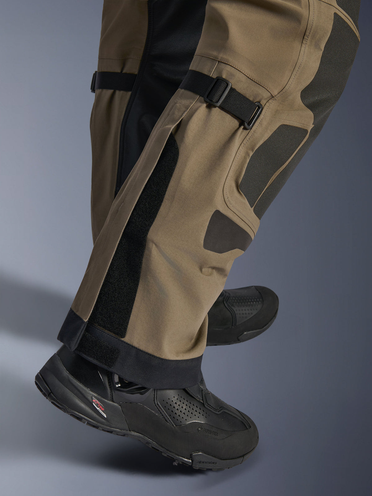 Close-up of Alpinestars Nazca 3L GORE-TEX PRO Pants in Walnut Black, showing technical details of the lower leg including calf volume adjusters and Superfabric reinforcements, worn with black adventure motorcycle boots.