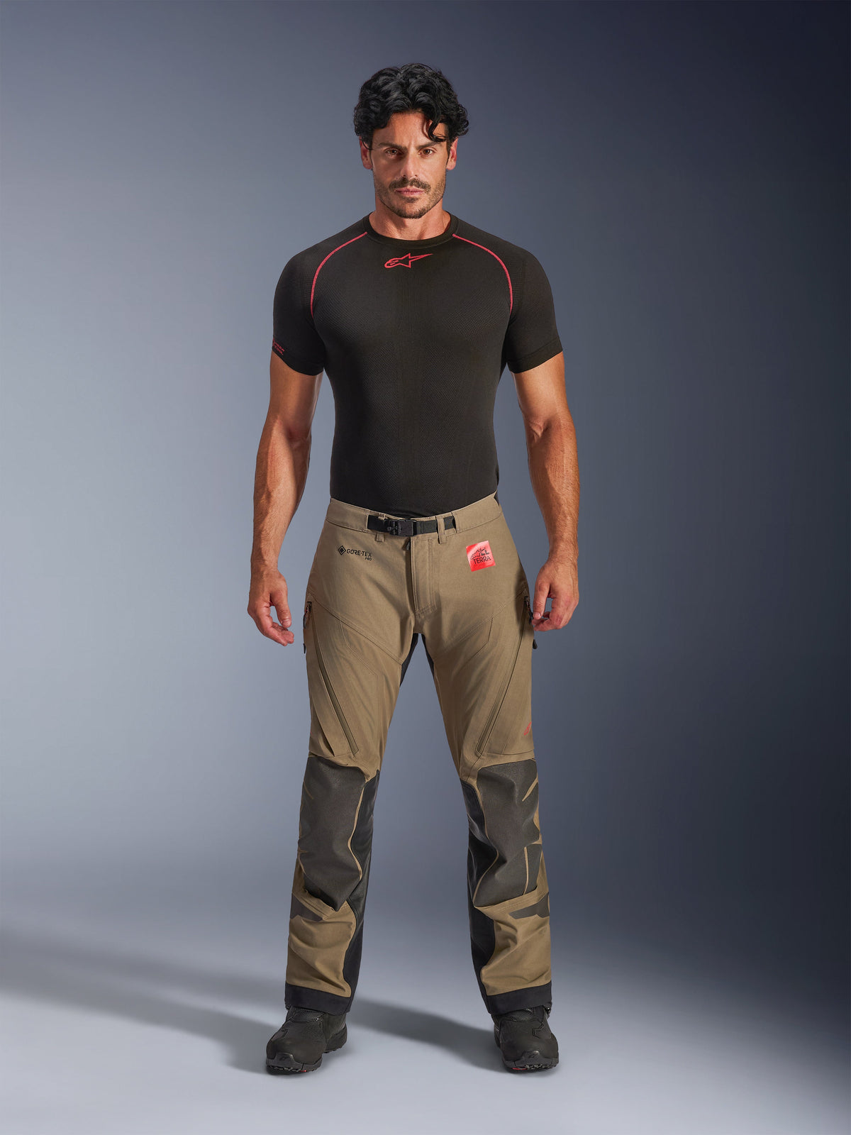 Male model wearing Alpinestars Nazca 3L GORE-TEX PRO Pants in walnut brown and black, technical adventure riding gear featuring Superfabric knee reinforcements and a magnetic waist buckle, paired with a black Alpinestars base layer shirt, studio front view.