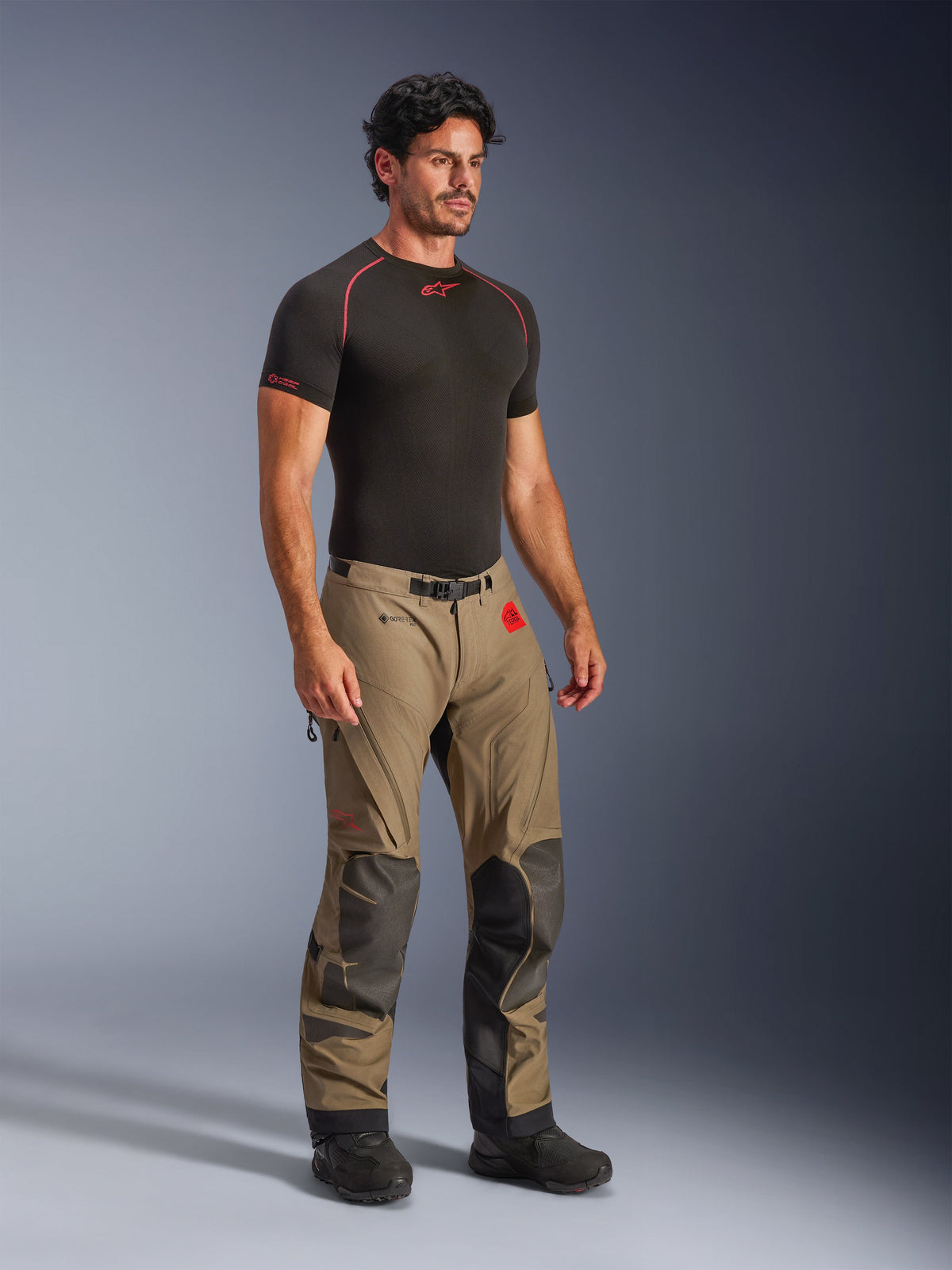 Alpinestars Nazca 3L GORE-TEX PRO Pants in Walnut Black, adventure motorcycle pants with black Superfabric reinforcements on knees, worn by a model with a black compression base layer, featuring a magnetic buckle belt and red All Terra branding.