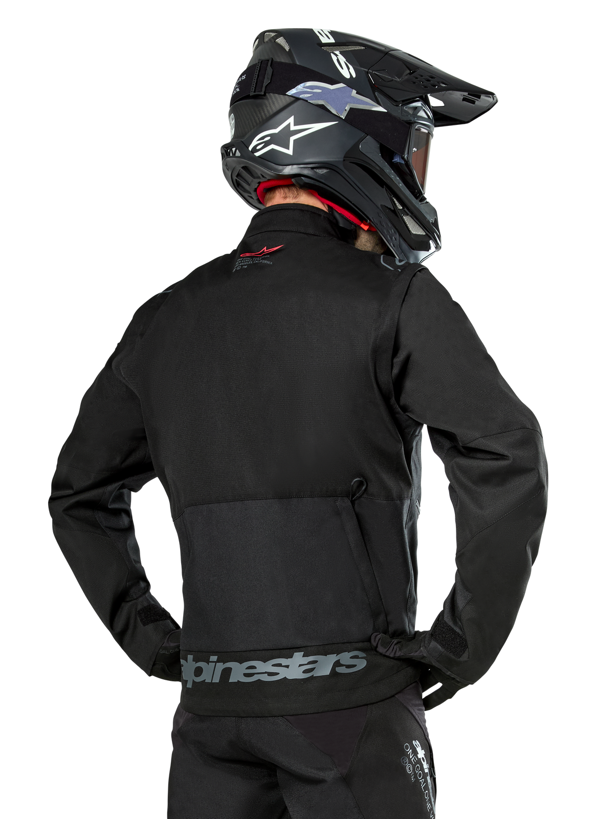 Rückansicht eines Fahrers in der Alpinestars Pro-Dura Jacke in Schwarz, strapazierfähige Enduro-Jacke aus Ripstop-Gewebe, mit einer großen Cargo-Tasche auf der Rückseite und grauem Alpinestars-Schriftzug am unteren Rücken, kombiniert mit einem schwarzen Motocross-Helm.