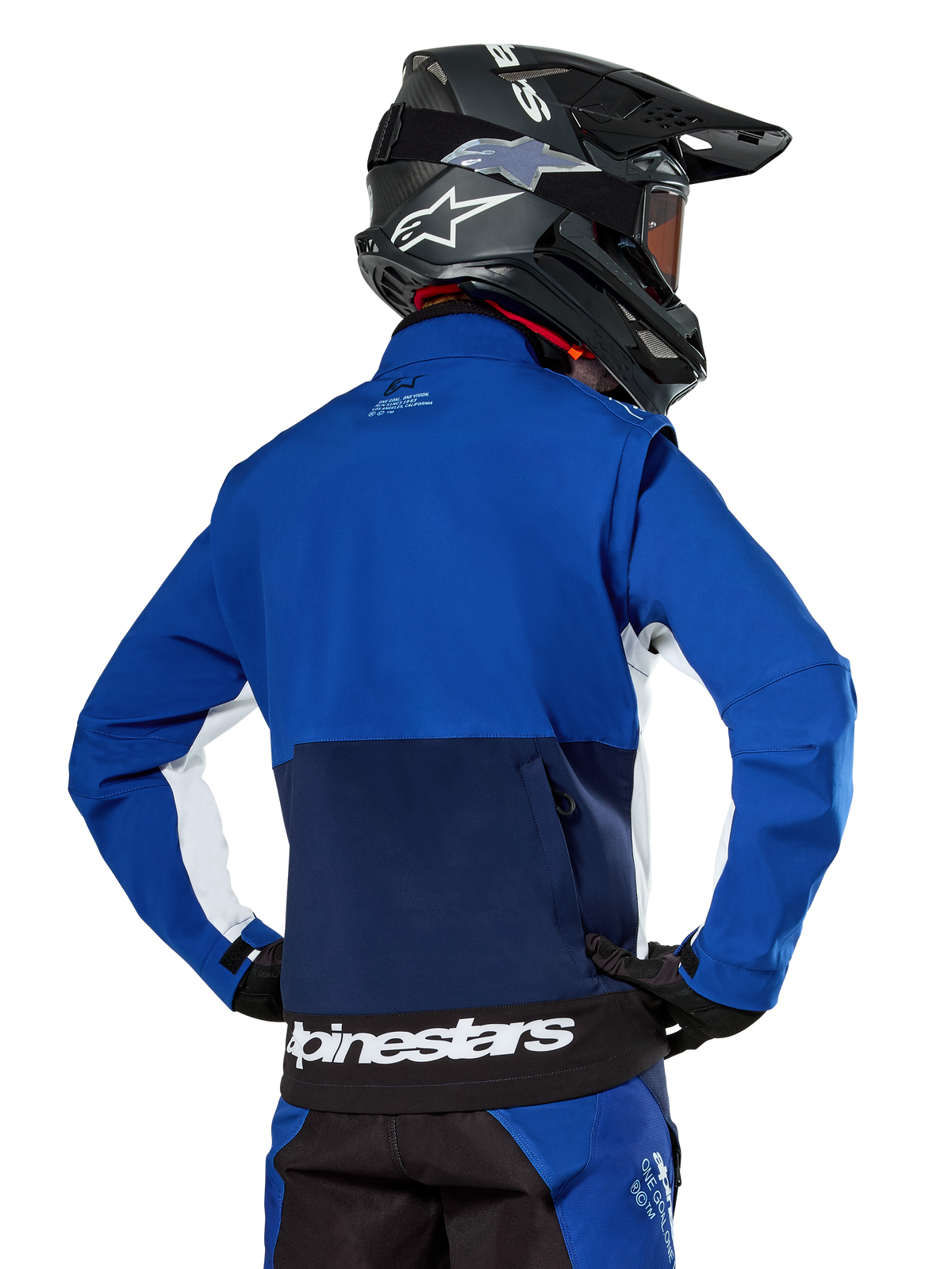 Alpinestars Lite-Dura Softshell-Jacke in Navy, Bright Blue und Weiß, Rückansicht mit großer Reißverschlusstasche zur Ärmelaufbewahrung, weißen Seitenakzenten und Alpinestars-Branding am Saum, getragen mit einem schwarzen Motocross-Helm und passender Ausrüstung.