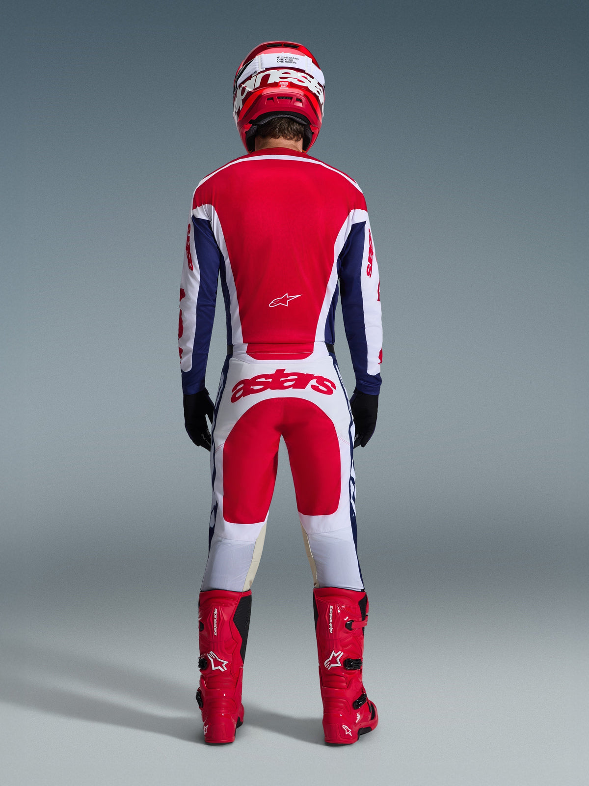 Motocross rider from behind wearing full racing gear, Alpinestars 2026 Racer Riway Pants in White Blue and Red with matching jersey, red boots and helmet, featuring large 'astars' logo on the seat, coordinated motocross outfit.