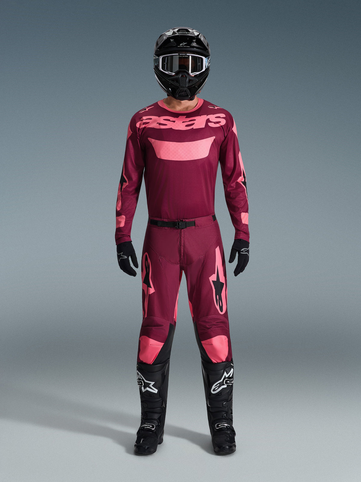 Motocross-Fahrer in kompletter Rennausrüstung, Trikot in Dark Red Coral mit rosa Akzenten, passende Racer Riway Hose, schwarzer Helm mit weißen Details, schwarze Stiefel mit weißen Logos, schwarze Handschuhe, abgestimmtes Outfit für den Offroad-Rennsport