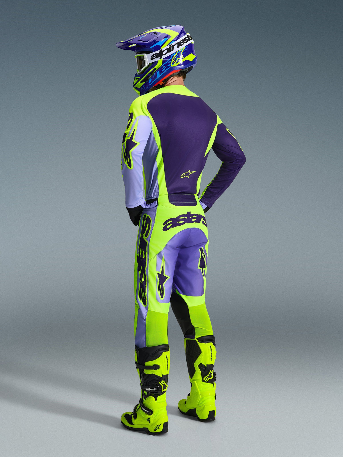 Motocross-Fahrer von hinten in der Alpinestars 2026 Racer Portl Hose in Purple Yellow Fluo, kombiniert mit einem passenden lila-gelben Trikot, Helm und leuchtend gelben Rennstiefeln, mit markantem 'astars'-Branding am Gesäß und Stern-Logos an den Beinen.
