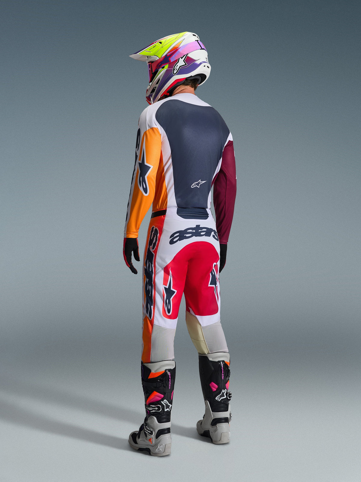 Motocross-Fahrer in der Rückansicht in kompletter Rennausrüstung, Alpinestars 2026 Racer Portl Hose in Rot, Hellgrau und Dunkelgrau, passendes Trikot mit orangefarbenen und weinroten Ärmeln, mehrfarbiger Grafikhelm, graue und schwarze Offroad-Stiefel mit pinkfarbenen Akzenten, professionelles Motocross-Outfit