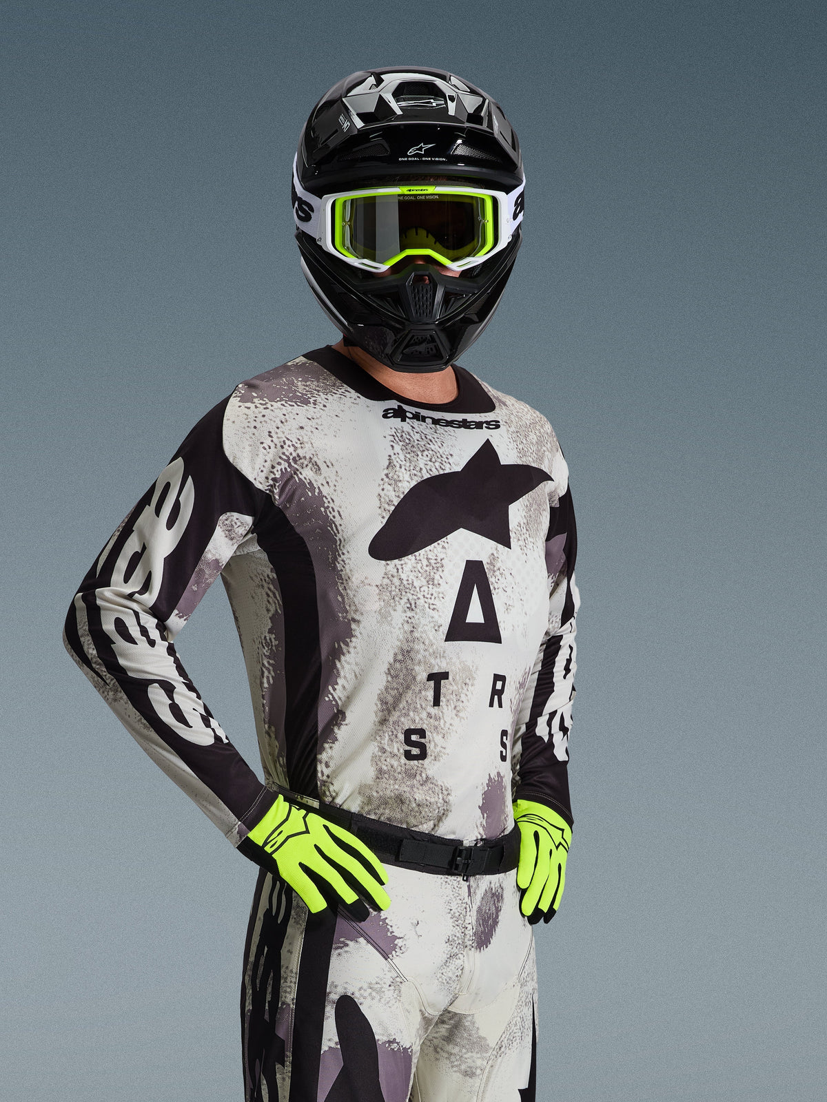 Alpinestars 2026 Racer Lahnd Trikot, Motocross-Trikot, Sand Camo mit grauem und schwarzem Muster, großes Astars-Branding auf der Brust, getragen mit schwarzem Helm, neongelber Brille und Handschuhen, Performance-Offroad-Rennausrüstung