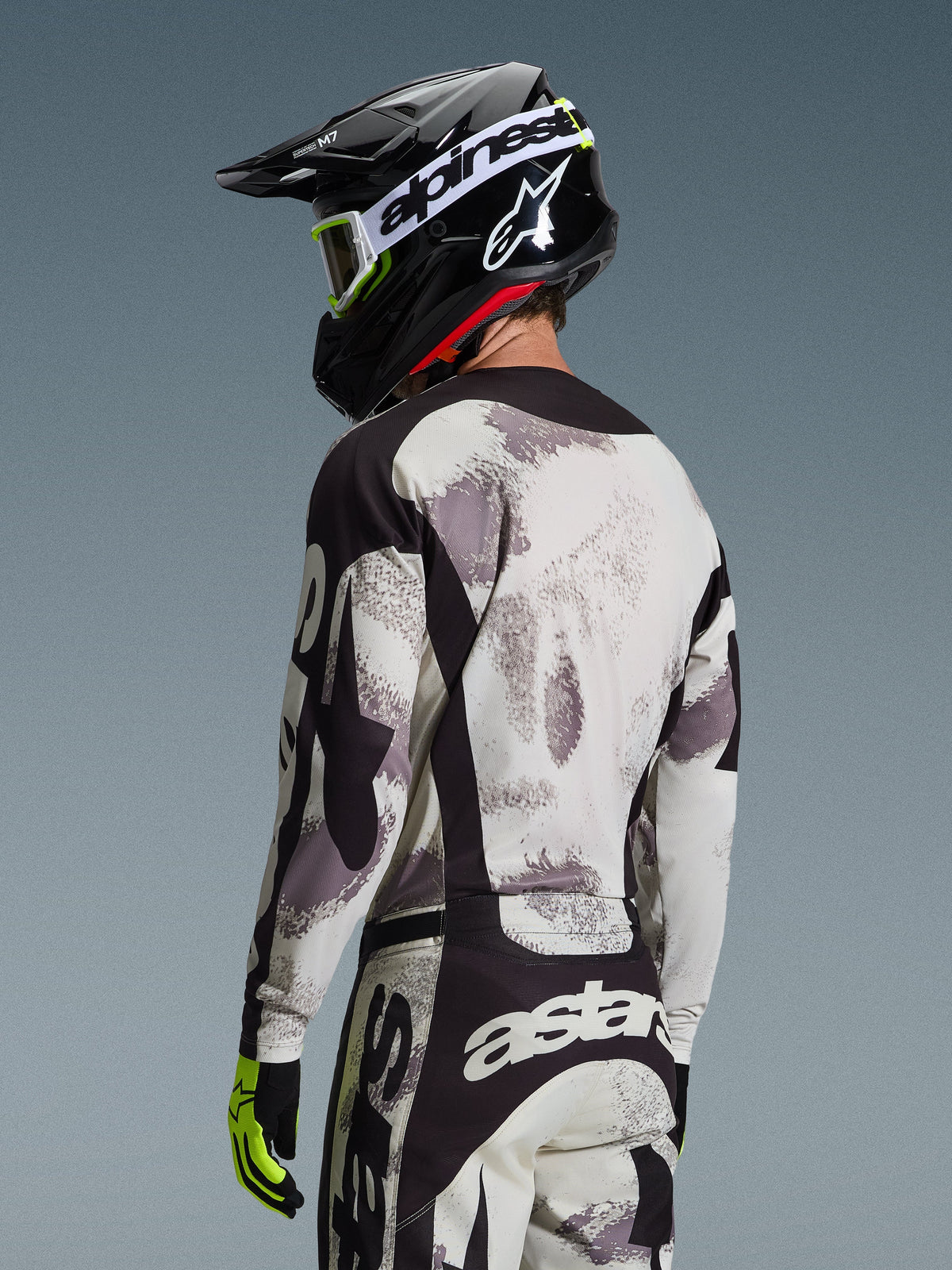 Motocross-Fahrer von hinten im 2026 Racer Lahnd Trikot in Sand Camo, grau und weiß abstrakt gemustertes Trikot mit schwarzen Akzenten, kombiniert mit schwarzem Alpinestars Helm, weißer Brille und neongelben Handschuhen, Rückansicht mit Fokus auf Performance-Passform für den Offroad-Rennsport.