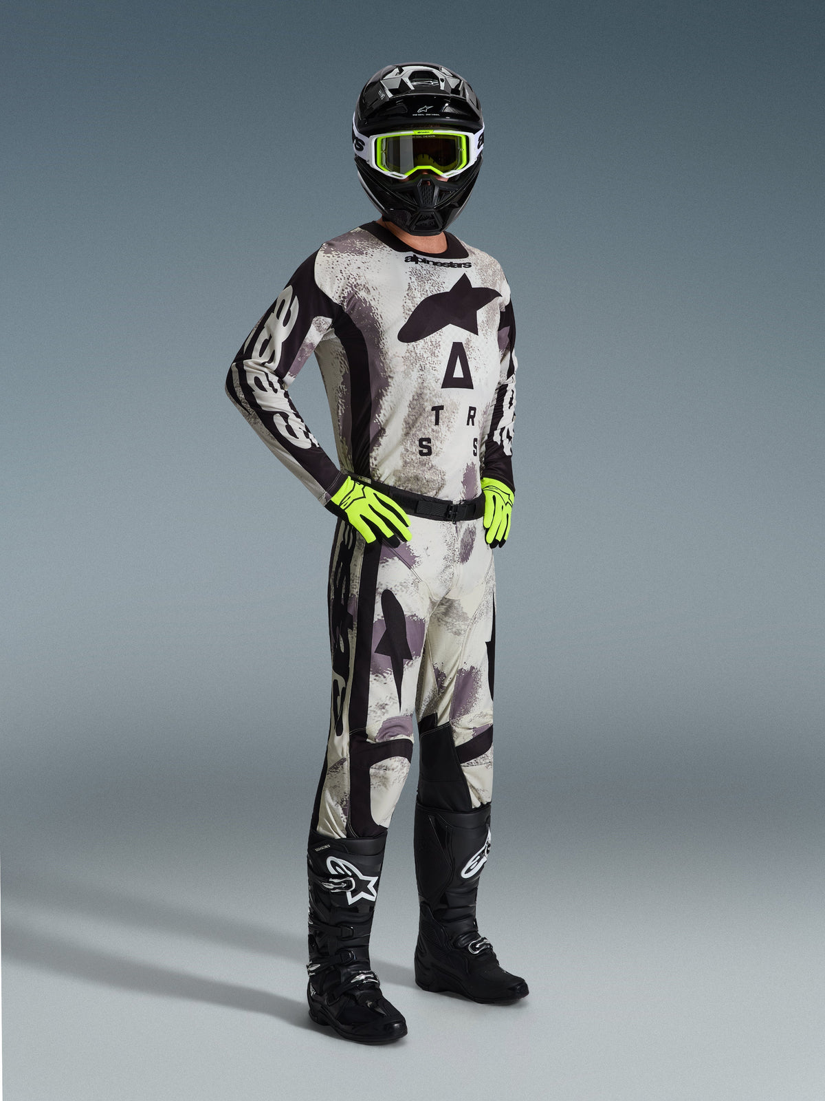 Motocross-Fahrer im 2026 Racer Lahnd Trikot in Sand Camo, grau-schwarzes Camouflage-Muster mit Alpinestars-Branding, passende Motocross-Hose, neongelbe Handschuhe, schwarzer Helm mit gelber Brille und schwarze Stiefel, komplette professionelle Offroad-Rennausrüstung.